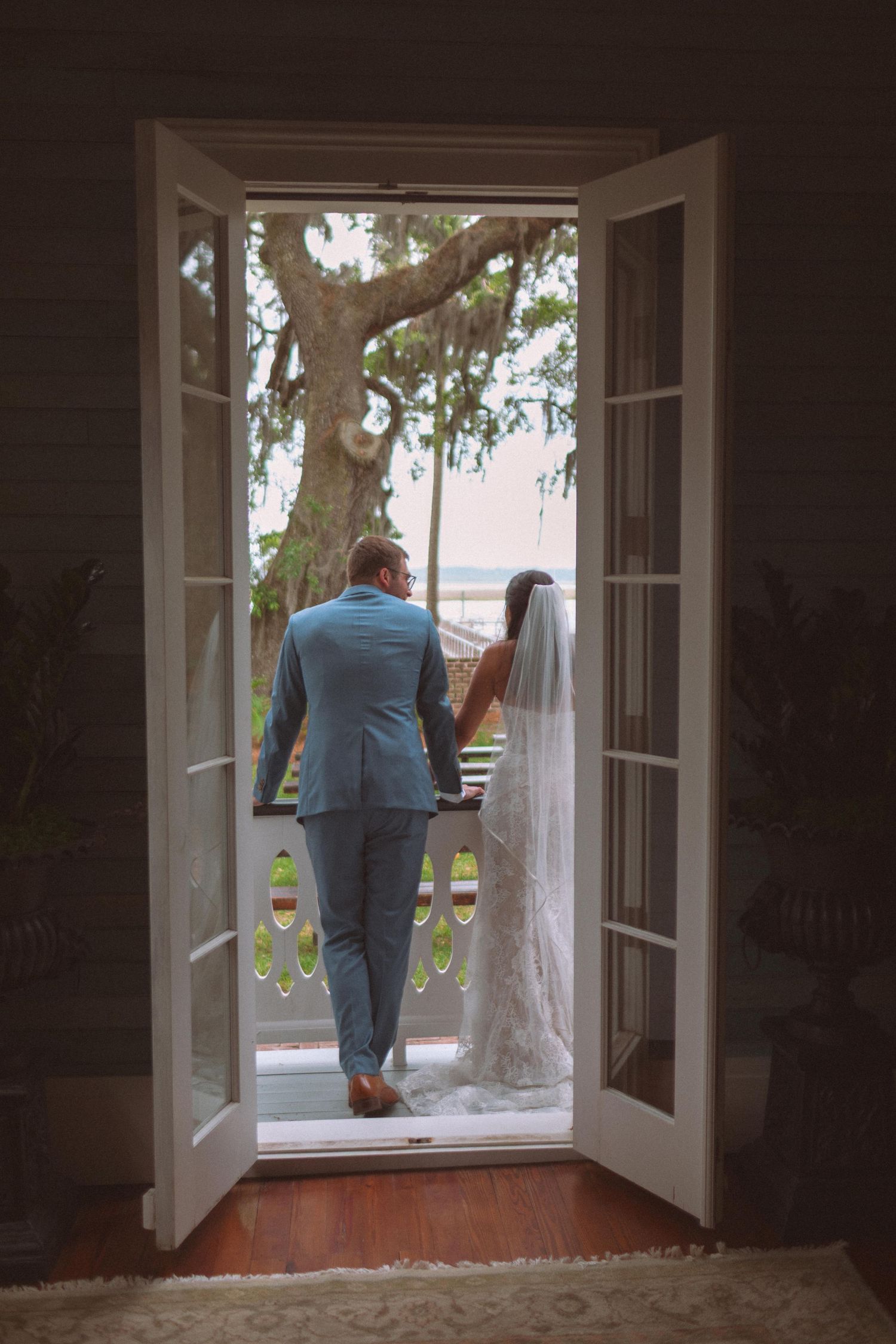 Bride and groom holding hands walking through French doors toward a scenic outdoor view on their wedding day.