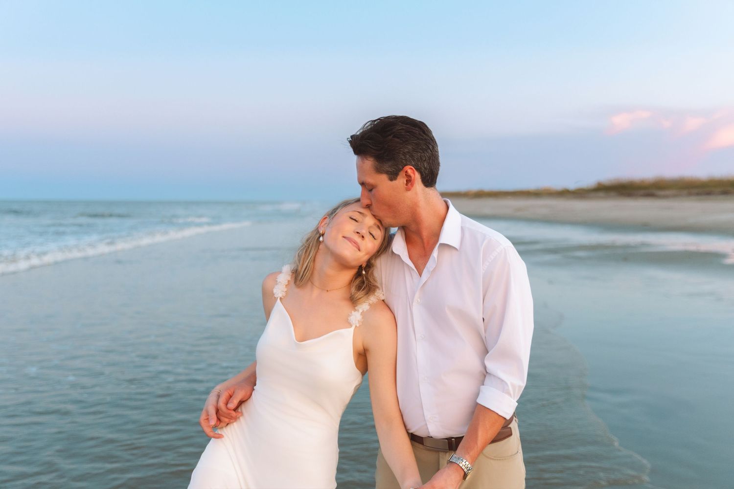 Couple on beach at dusk, man kissing womans cheek, woman in white slip dress, pastel sky background.