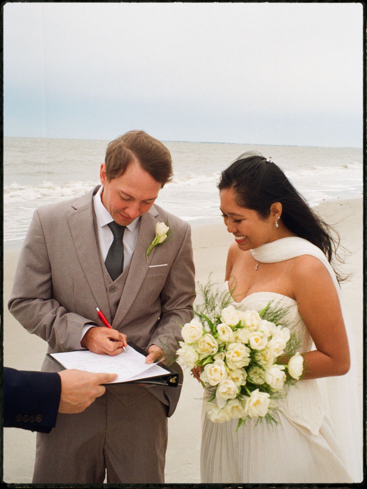 Groom in a tan suit signing wedding documents on a beach while bride holds white roses bouquet nearby.