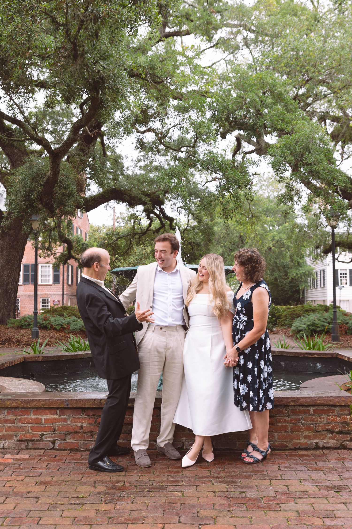 Bride and groom with family gathered around a fountain in a charming outdoor square with lush trees.