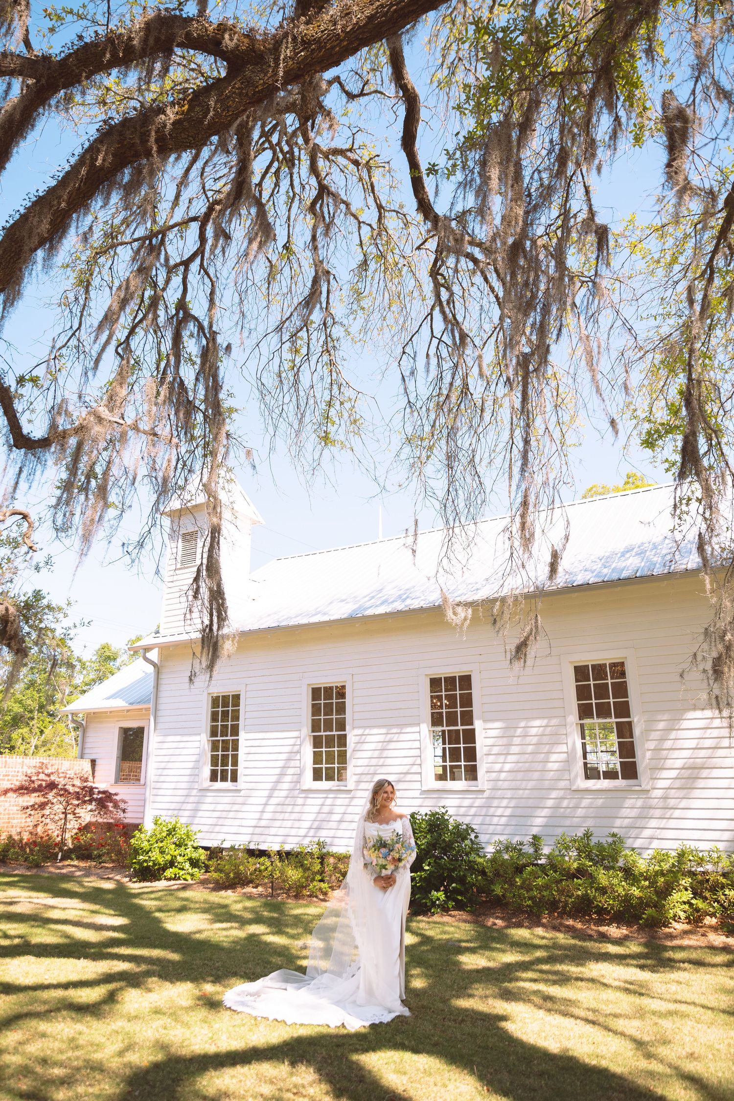 Bride in white gown poses before historic white chapel with Spanish moss-draped oak tree.