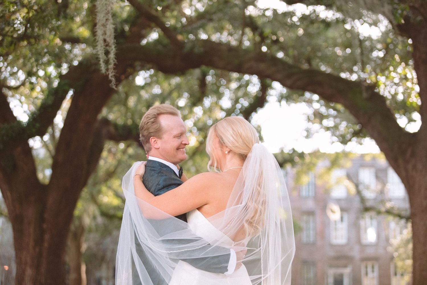 Groom lifts smiling bride in wedding veil beneath a large oak tree outside a historic building.