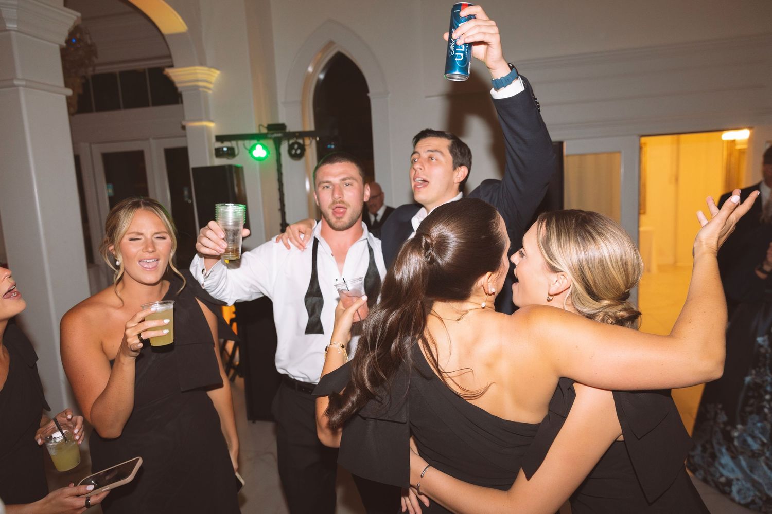 Wedding guests dancing and raising drinks in a elegant white venue during a lively reception celebration.