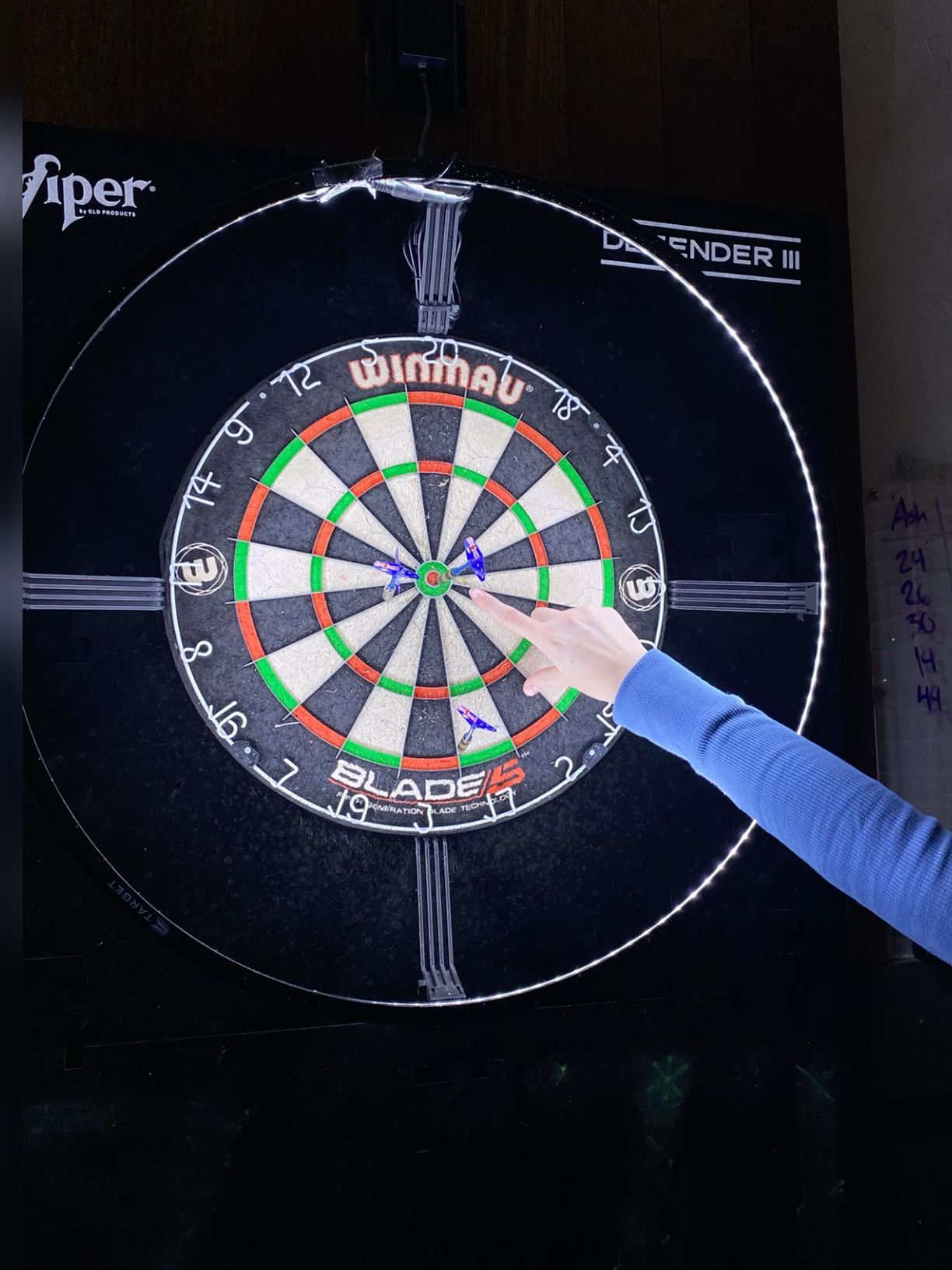 Person pointing at darts clustered near the bullseye on a Winmau Blade dartboard.