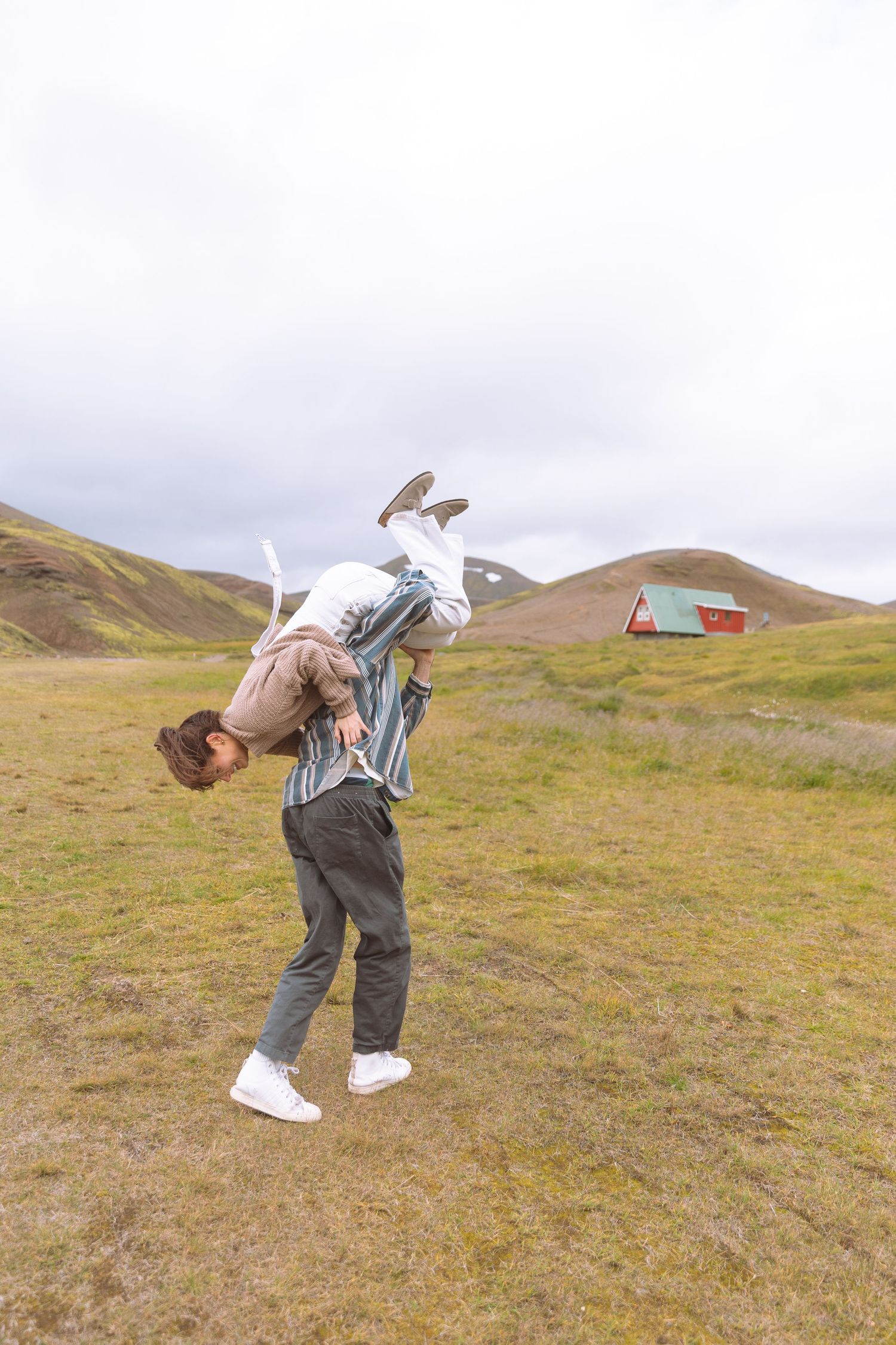 Playful couple outdoors, man carrying woman over his shoulder in a green hilly landscape.