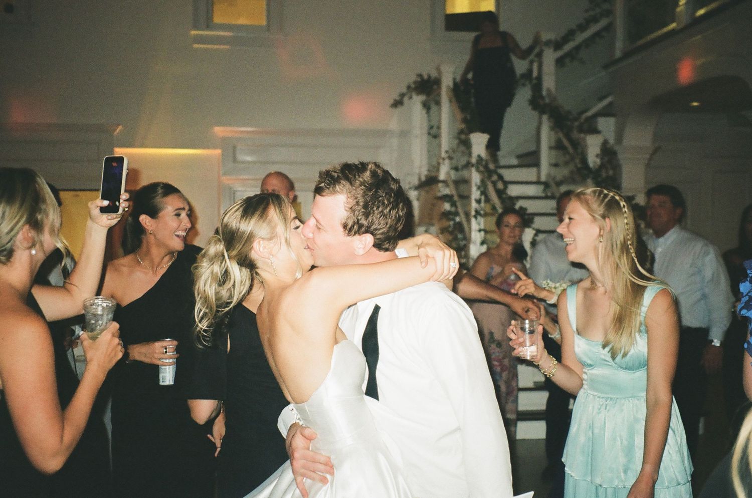 Bride and groom share a romantic kiss on the dance floor surrounded by guests at an elegant wedding reception.