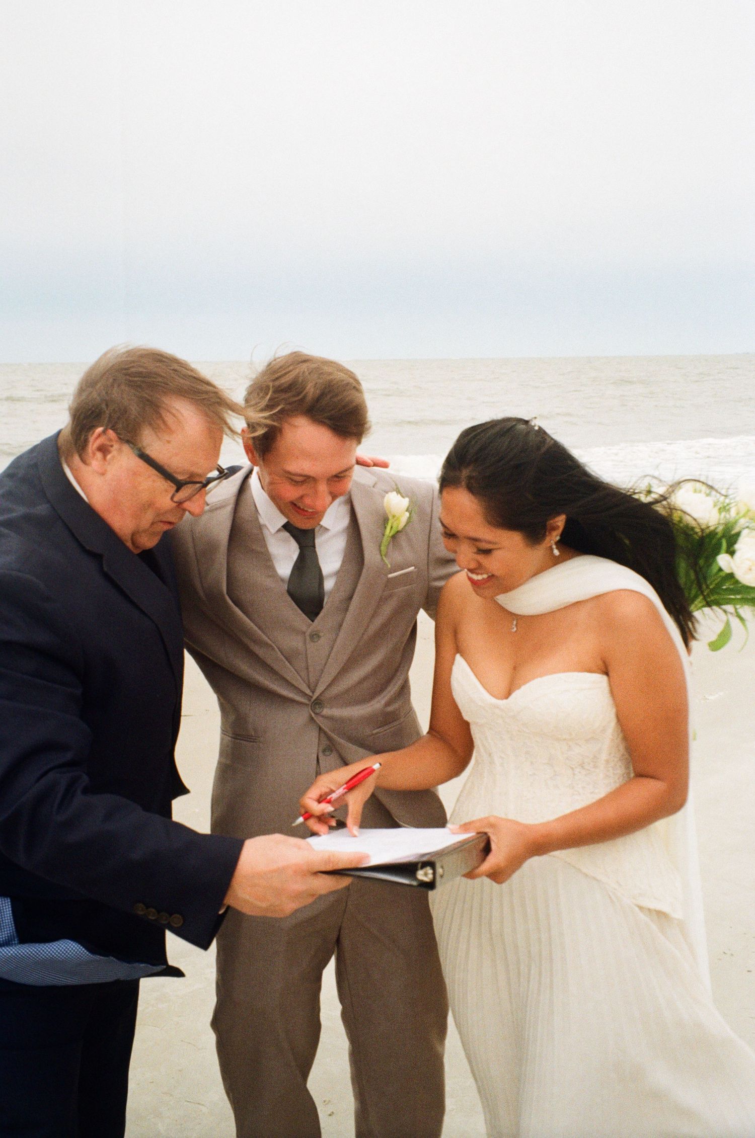 Bride and groom signing marriage certificate with officiant during a romantic beach wedding ceremony.