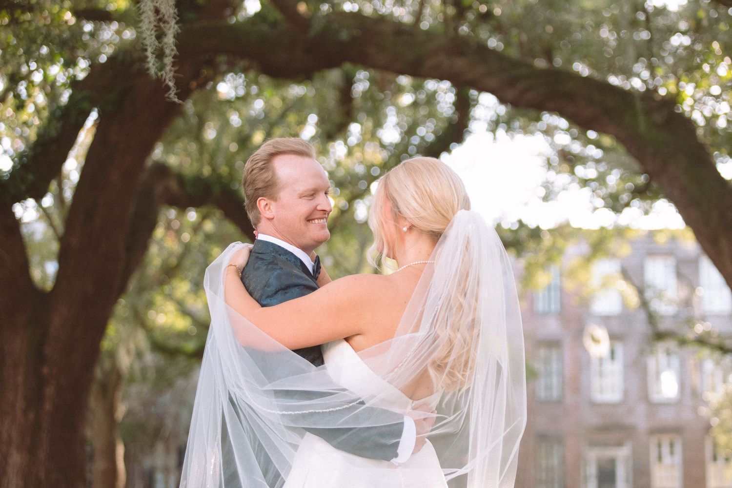 Smiling bride and groom embracing under moss-draped oak trees on their wedding day.