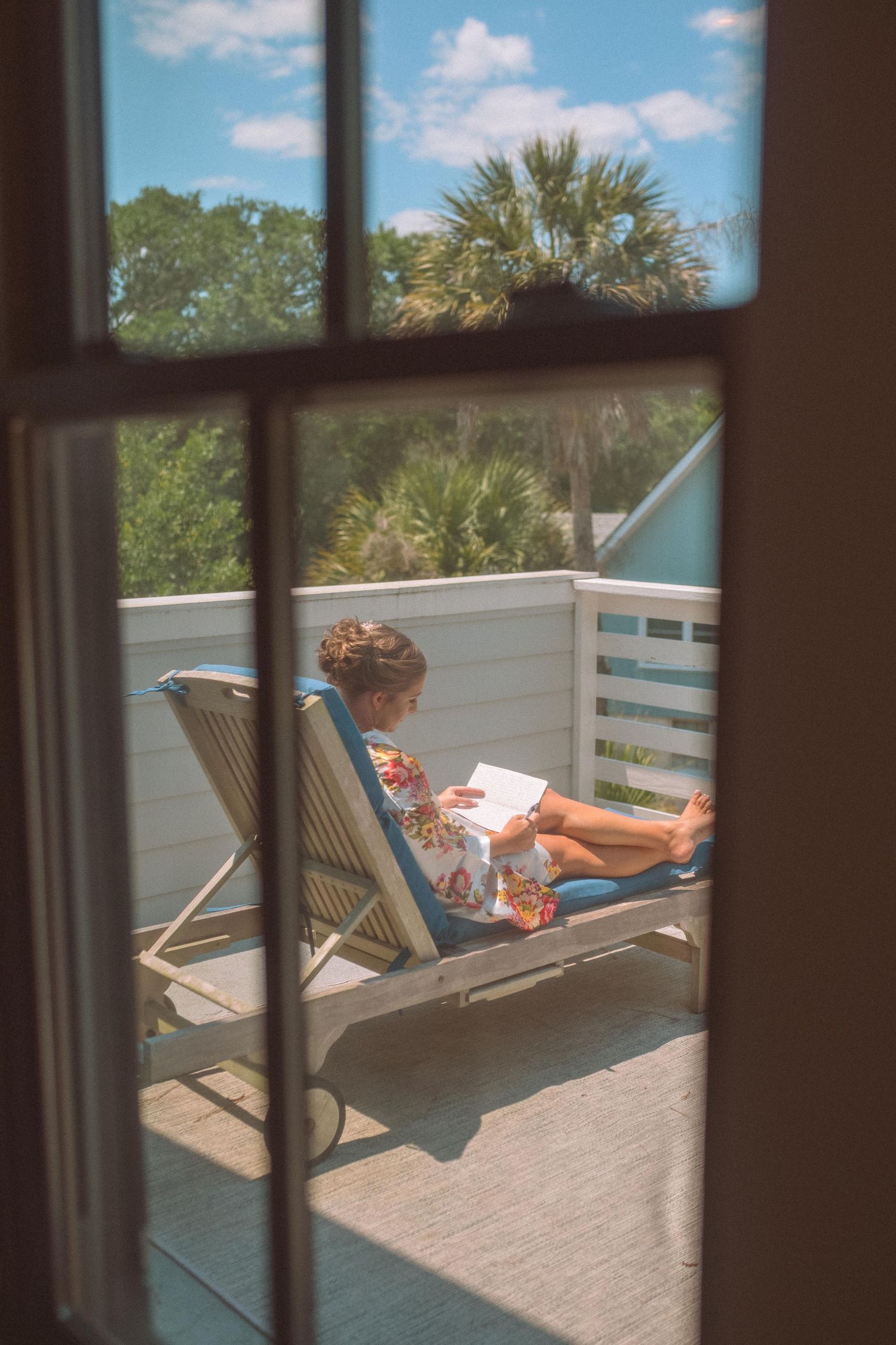 Woman relaxing on lounge chair reading book on sunny outdoor deck viewed through window.