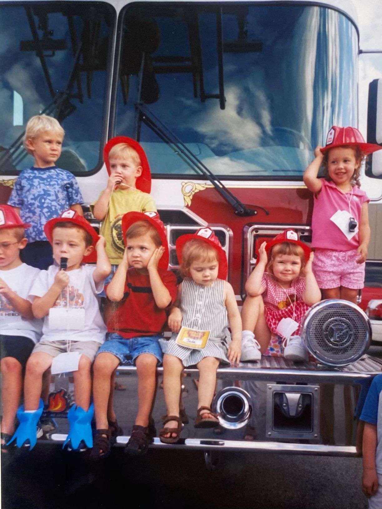Young children wearing red firefighter helmets sitting on front of a fire truck.