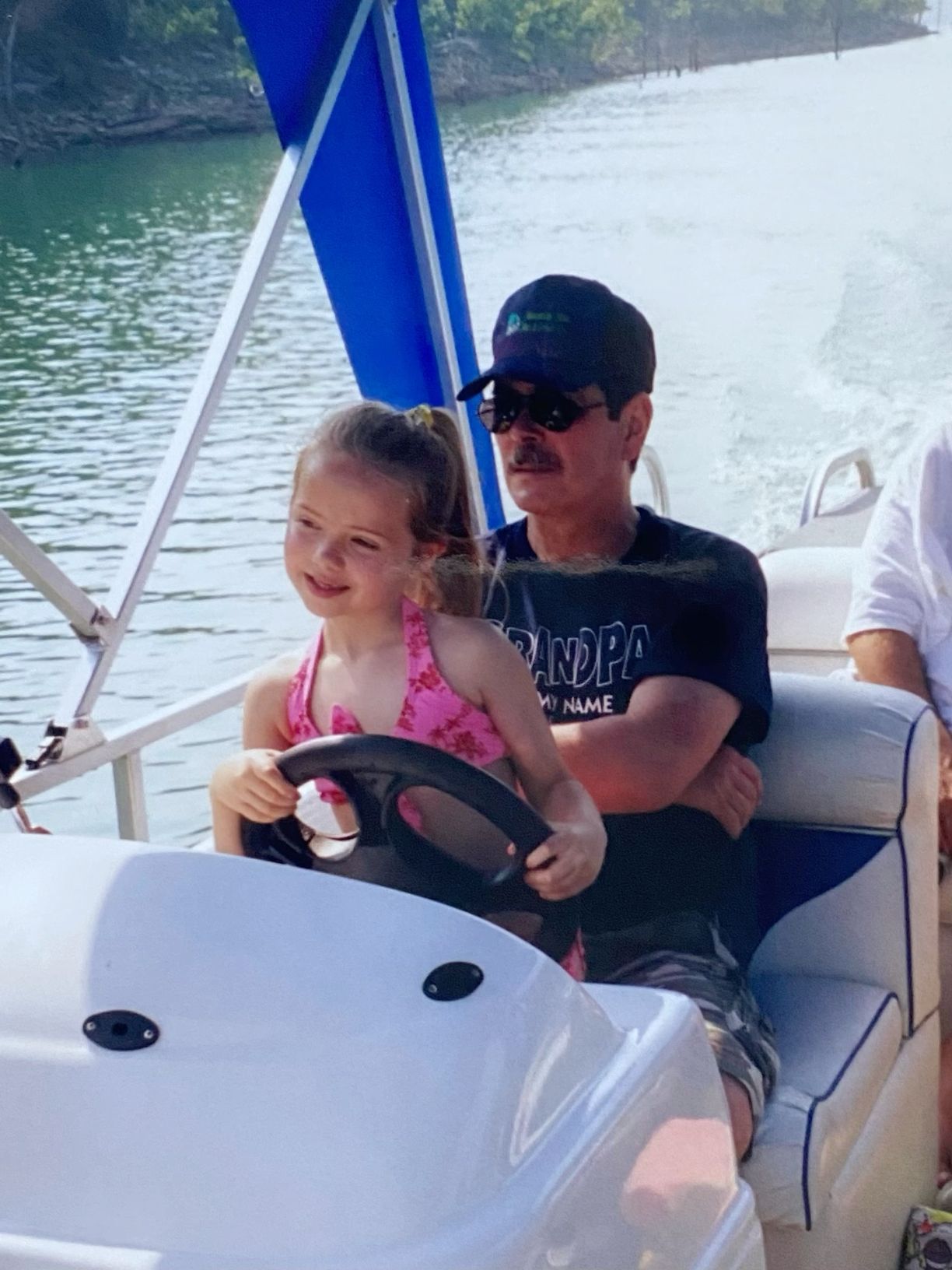 Young girl steering a pontoon boat with grandfather on a scenic lake.