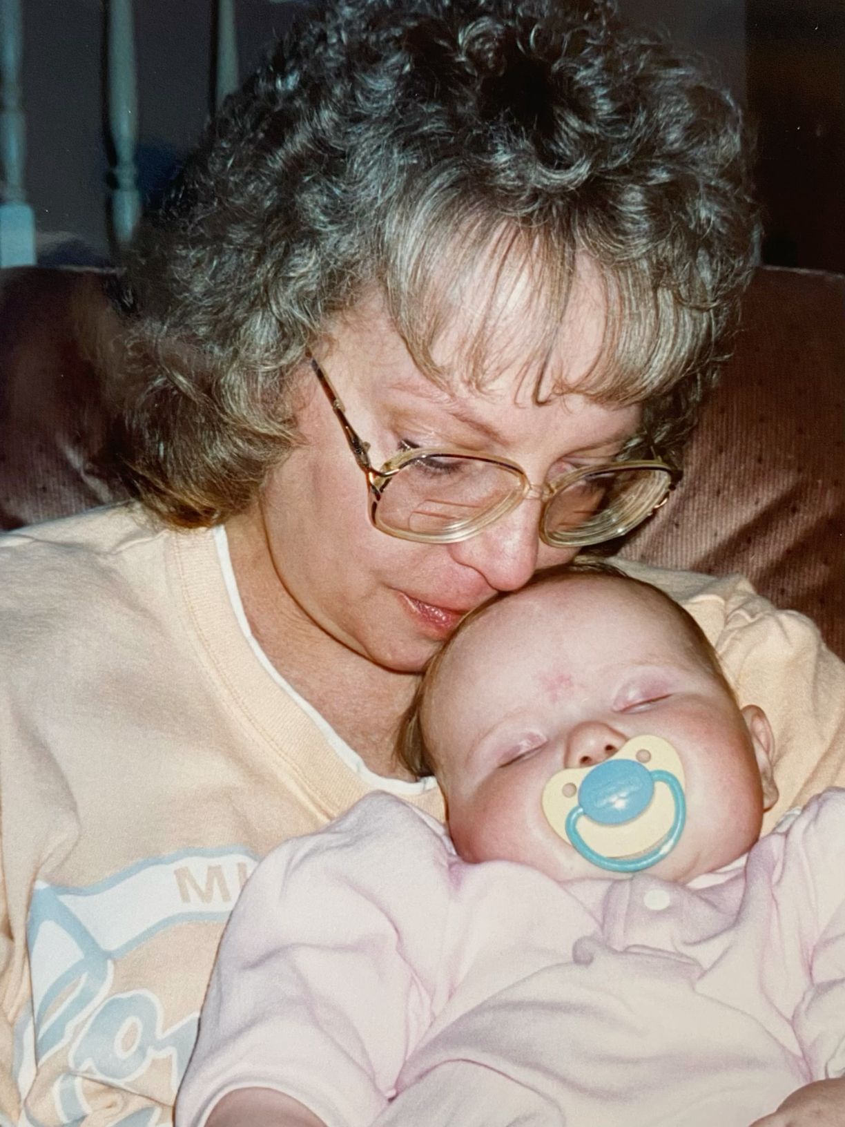 Grandmother with curly hair and glasses tenderly holding a sleeping newborn with a pacifier.