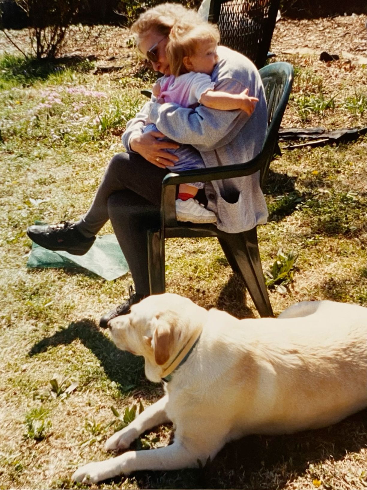 A woman cuddles a toddler in a garden chair while a yellow Labrador rests nearby.