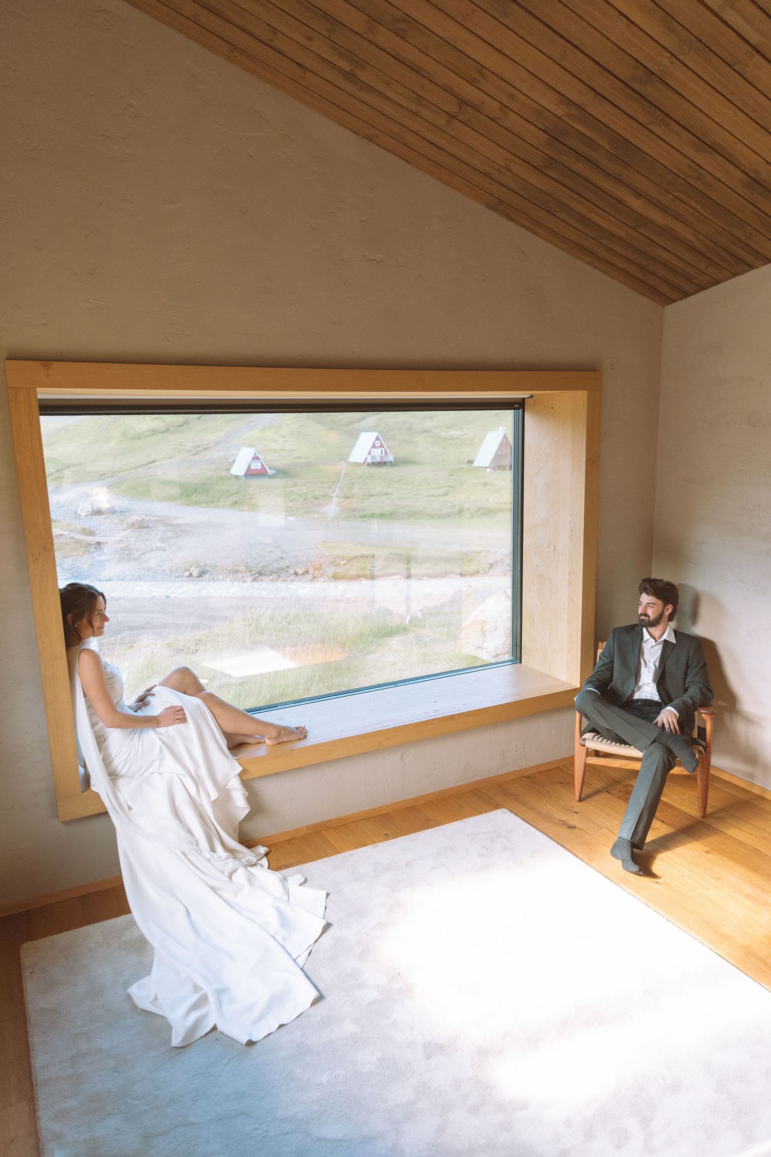 Bride and groom share a quiet moment in a modern wooden cabin with a large scenic window in Iceland.