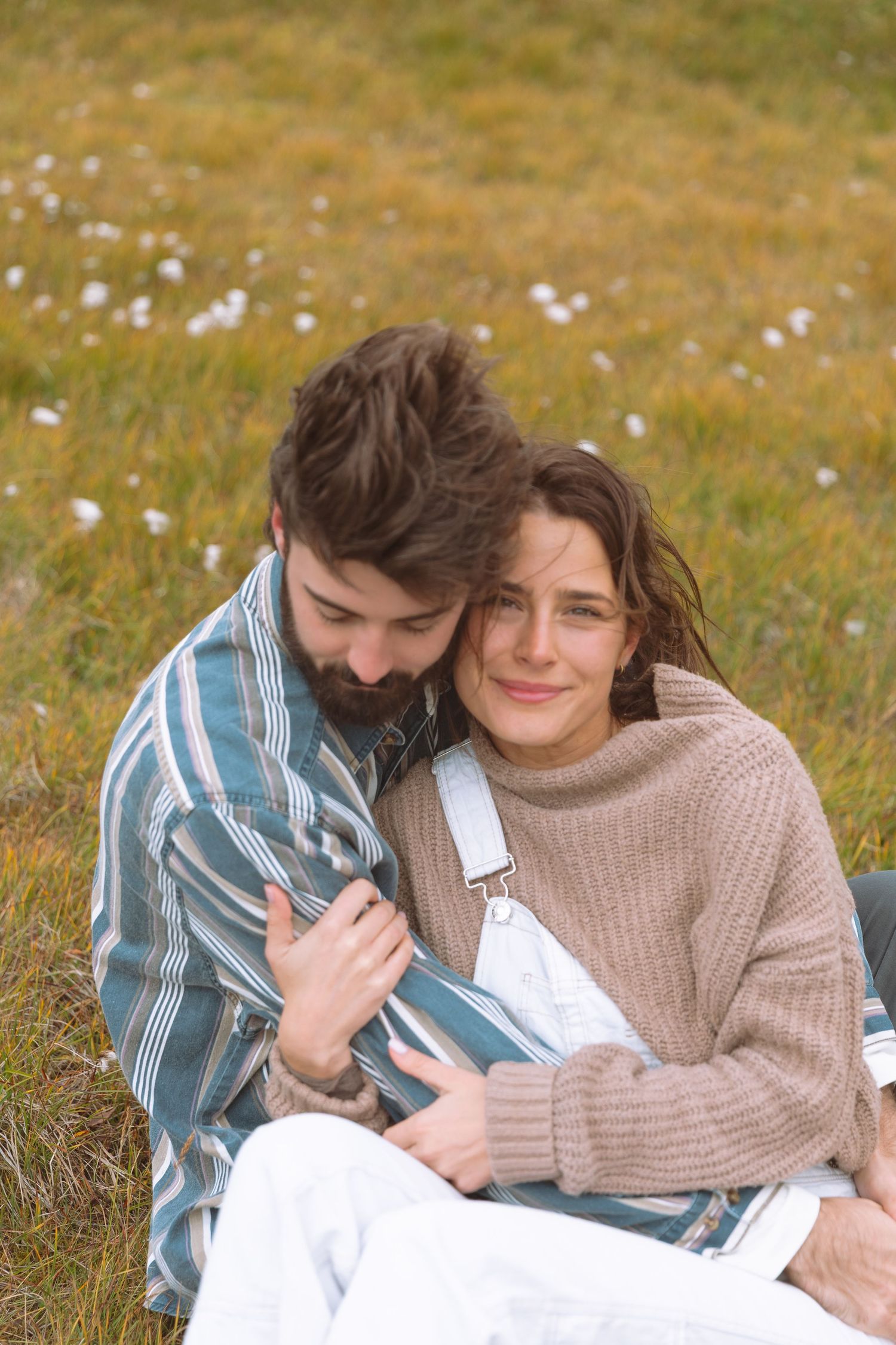 Bearded man hugging smiling woman in knit sweater while sitting on grass field.