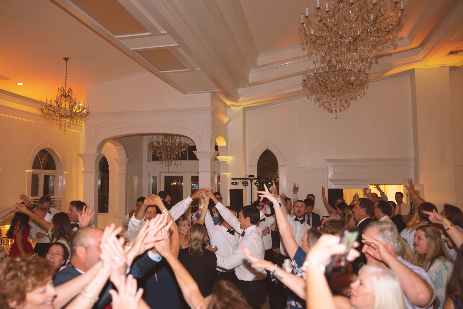 Elegant wedding reception dance floor with guests celebrating under crystal chandeliers in a grand white ballroom.