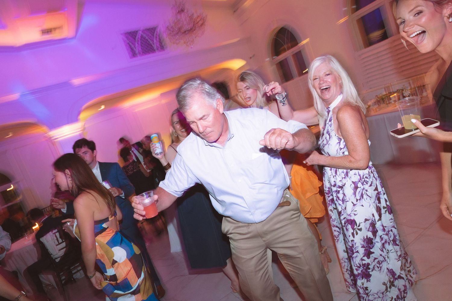 Guests enjoy lively dancing at a wedding reception with purple ambient lighting.