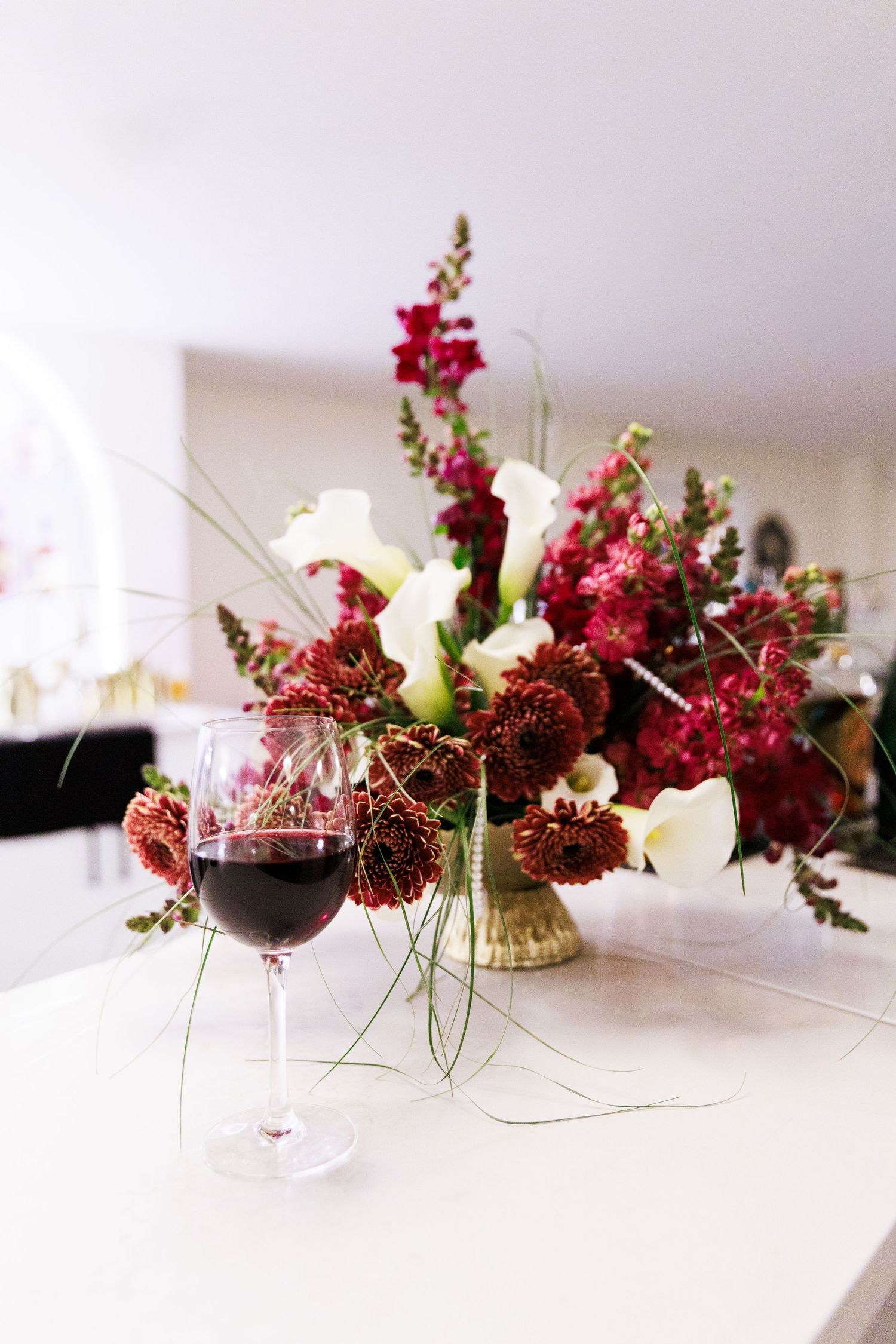 Elegant glass of red wine beside a lush floral arrangement with burgundy and white blooms on a white table.
