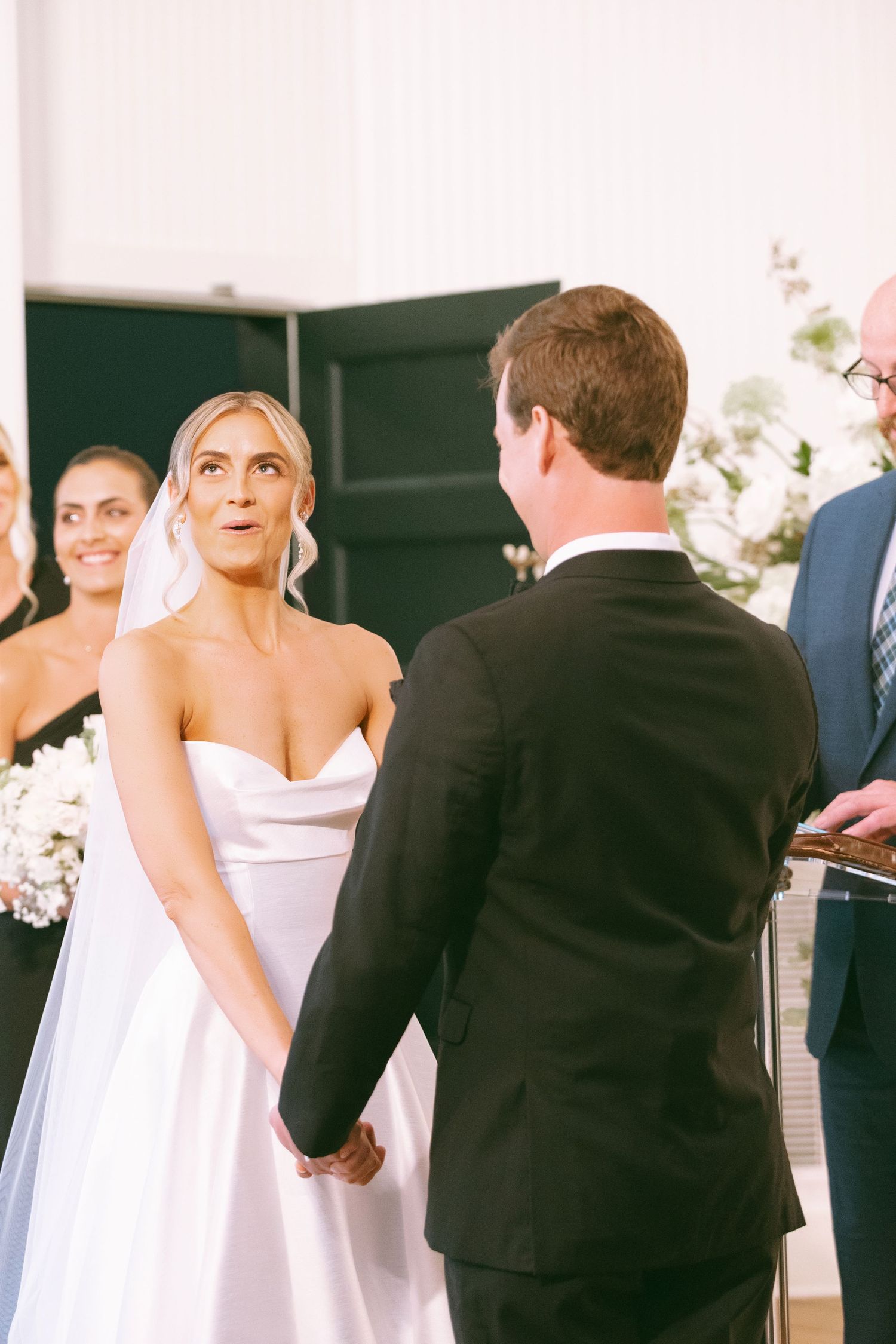 Bride and groom hold hands at wedding altar, bride in white satin gown with veil, groom in black suit.