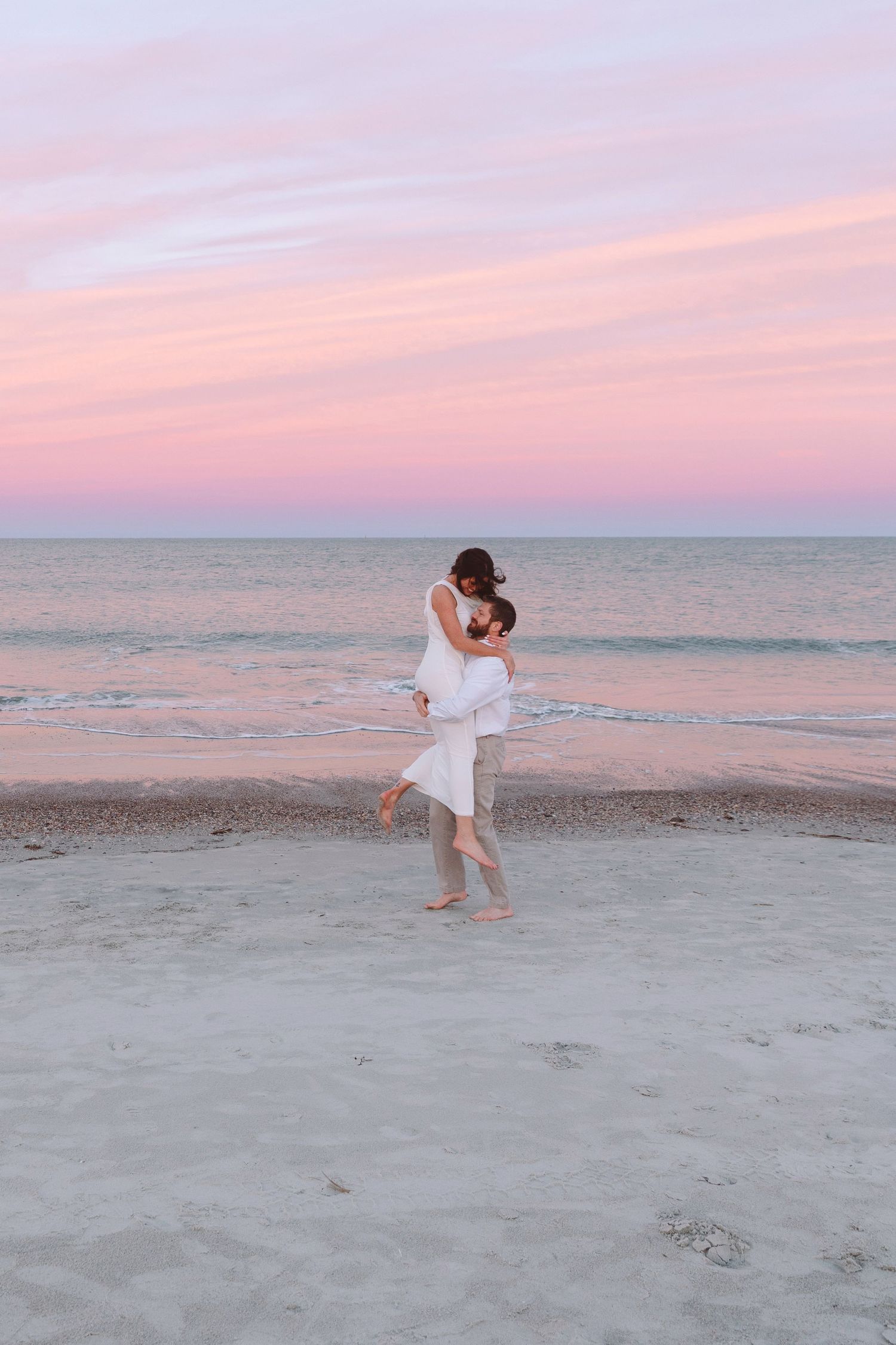 Couple embracing on a sandy beach at sunset with pink and purple sky over calm ocean waters.