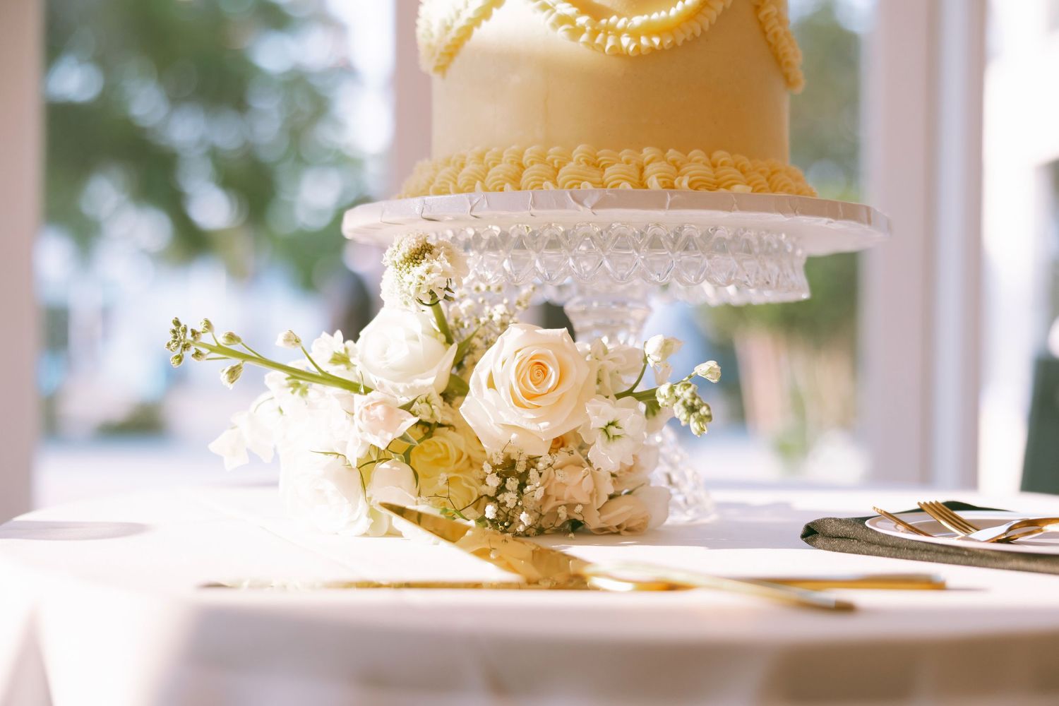Elegant two-tier wedding cake with yellow pearl details beside white roses bouquet on draped table.