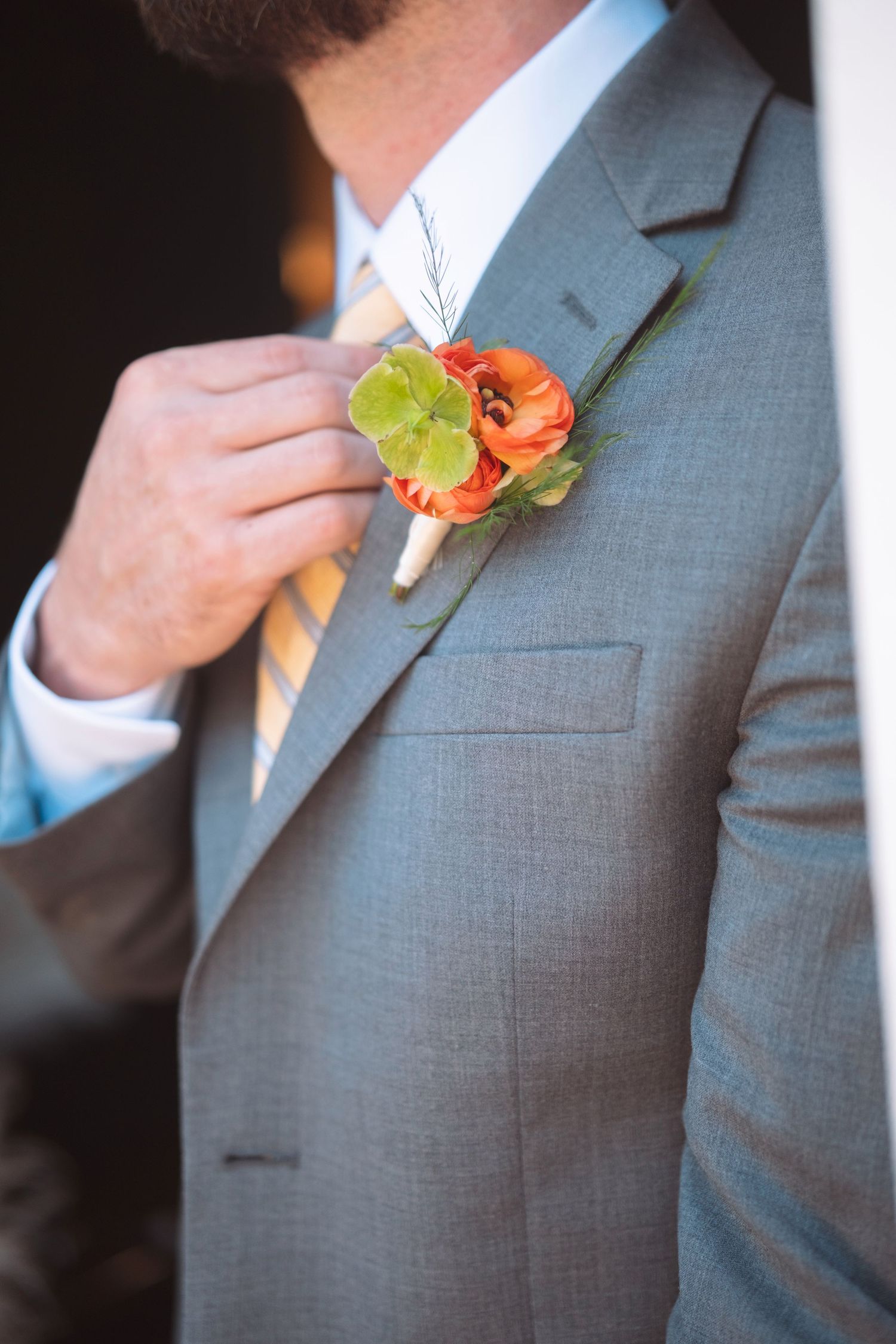 Groom in gray suit adjusting orange ranunculus boutonniere with yellow striped tie.