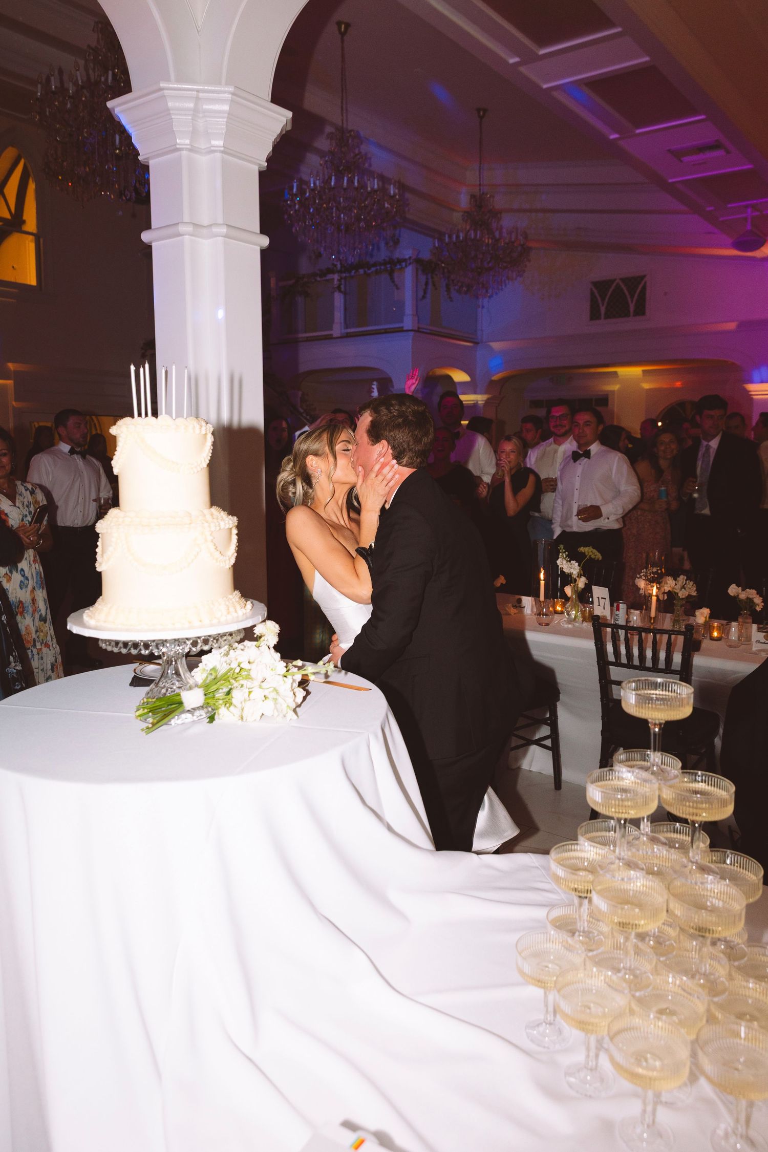 Bride and groom sharing a kiss beside white tiered wedding cake and champagne tower at reception.