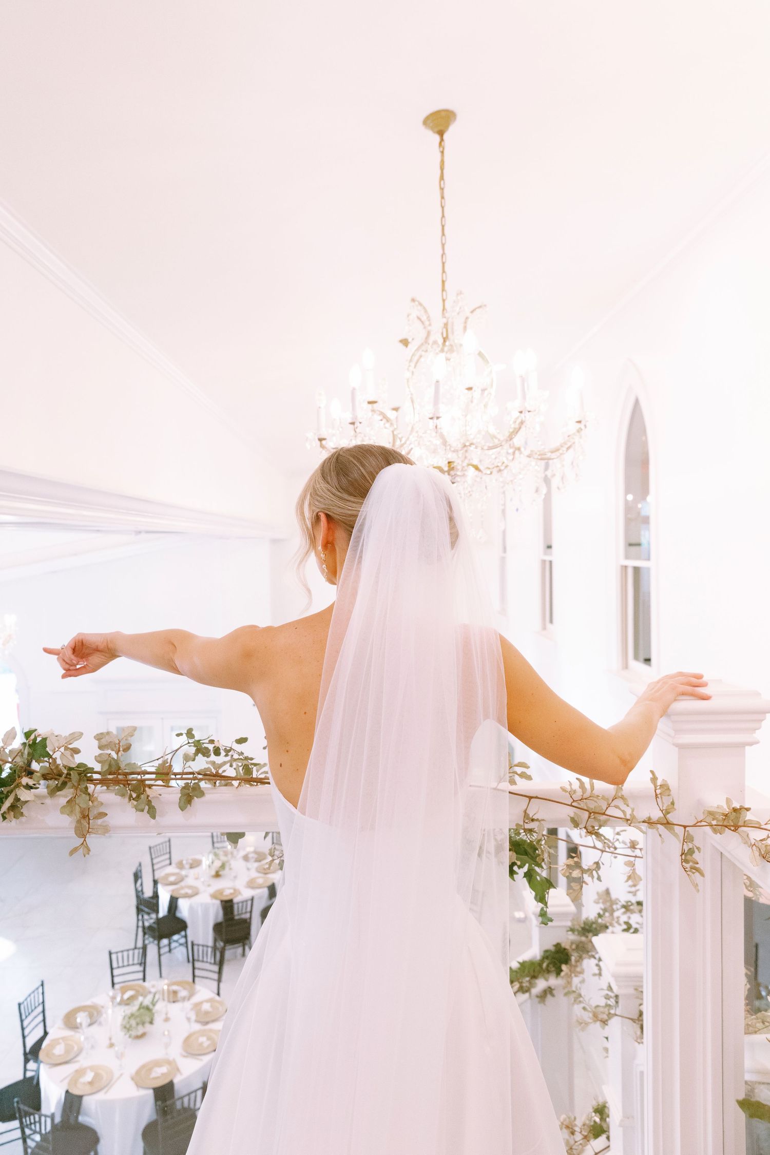 Bride in white gown and veil stands on balcony with arms outstretched overlooking elegant wedding reception hall with chandelier.