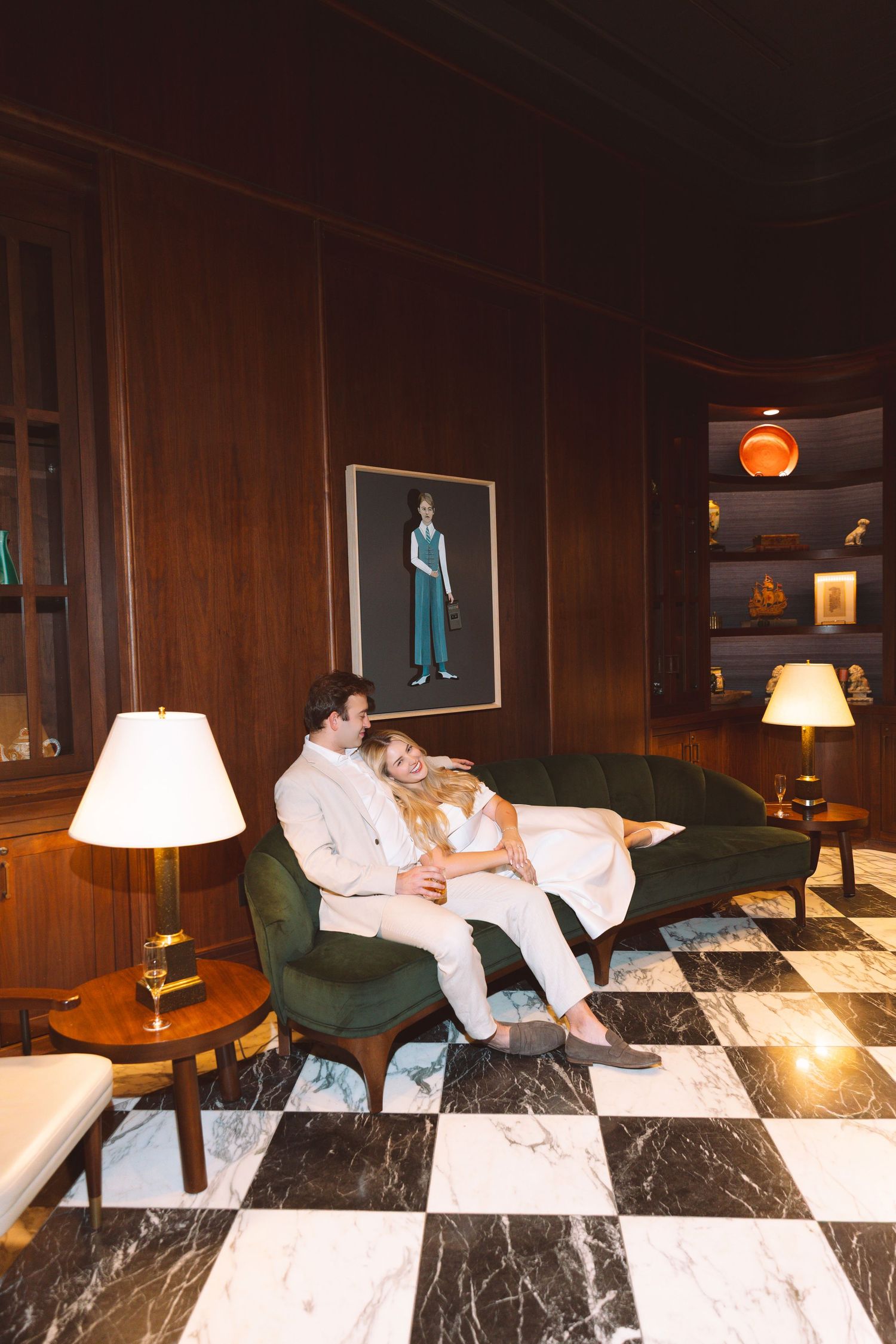 Couple relaxing together on a green velvet sofa in an elegant wood-paneled room with checkered marble floors.
