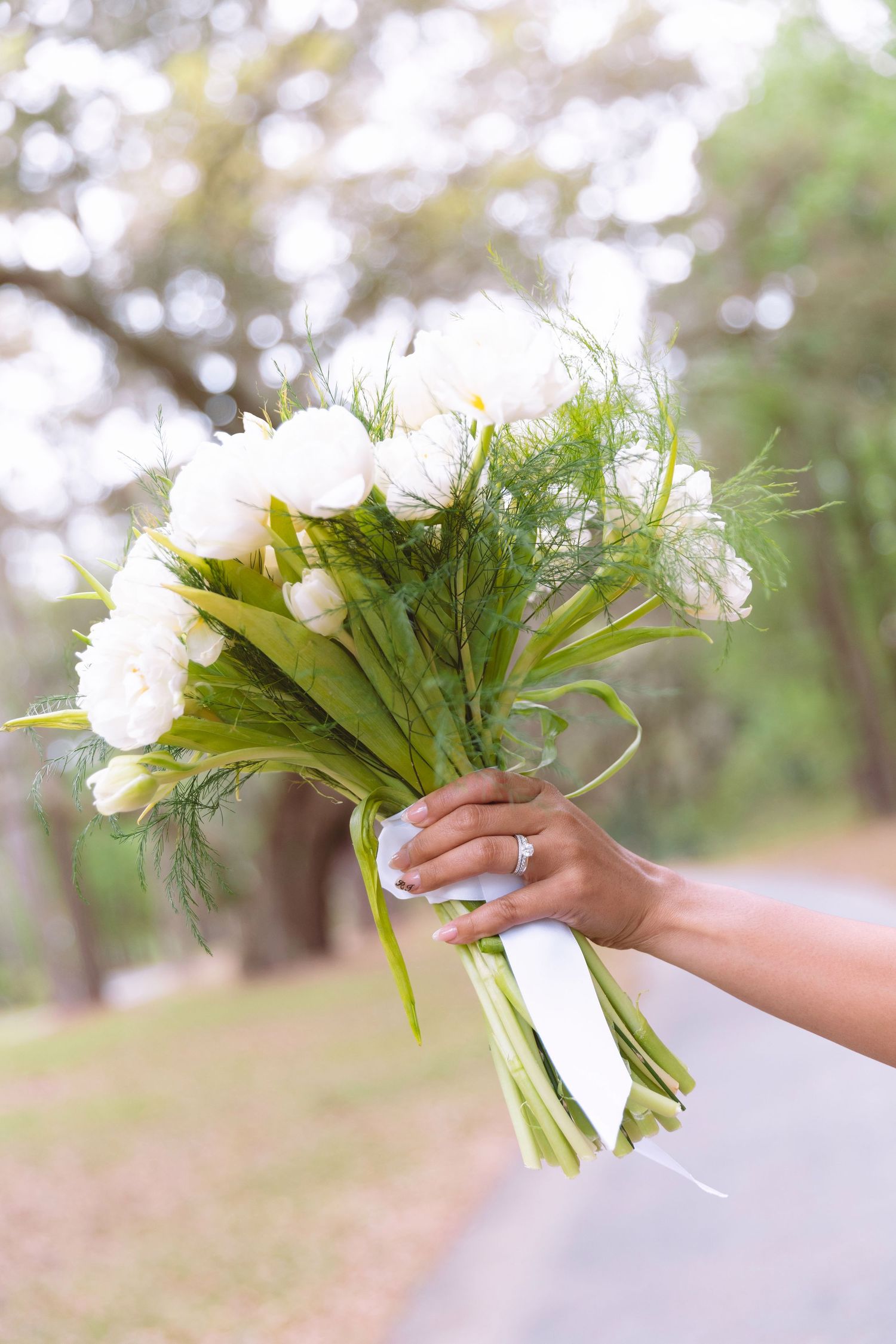 Bride holding a stunning white ranunculus wedding bouquet with green foliage tied with white ribbon.