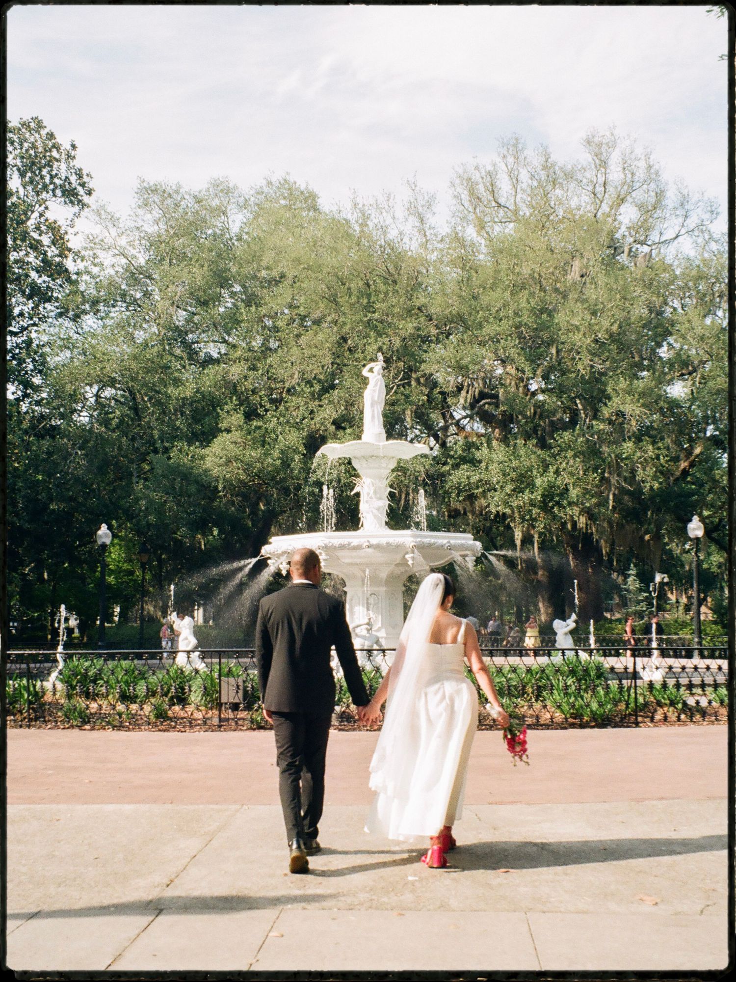 Bride and groom walk hand-in-hand toward ornate white fountain in scenic park setting.
