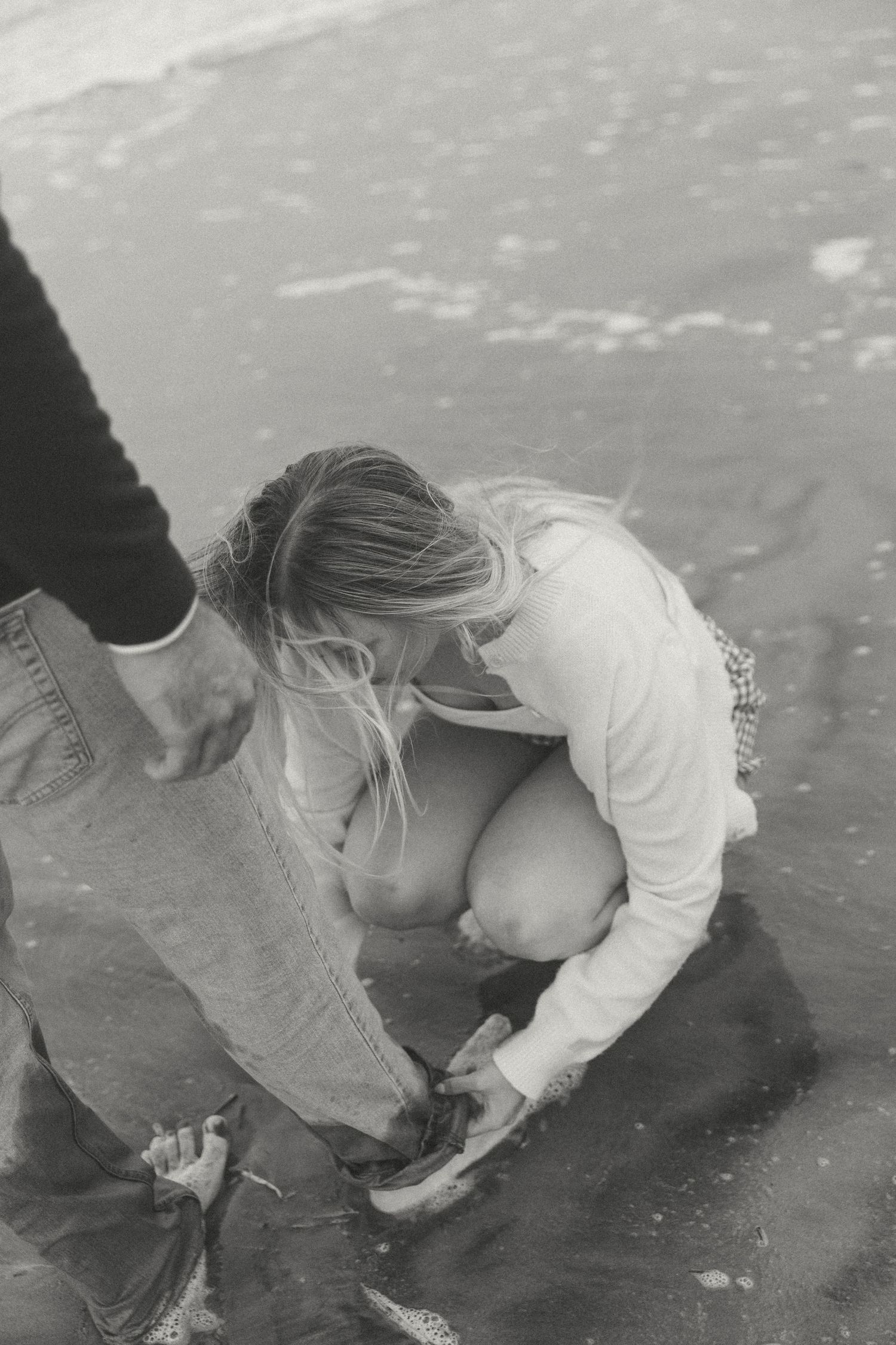 Black and white photo of woman crouching by water at the beach.