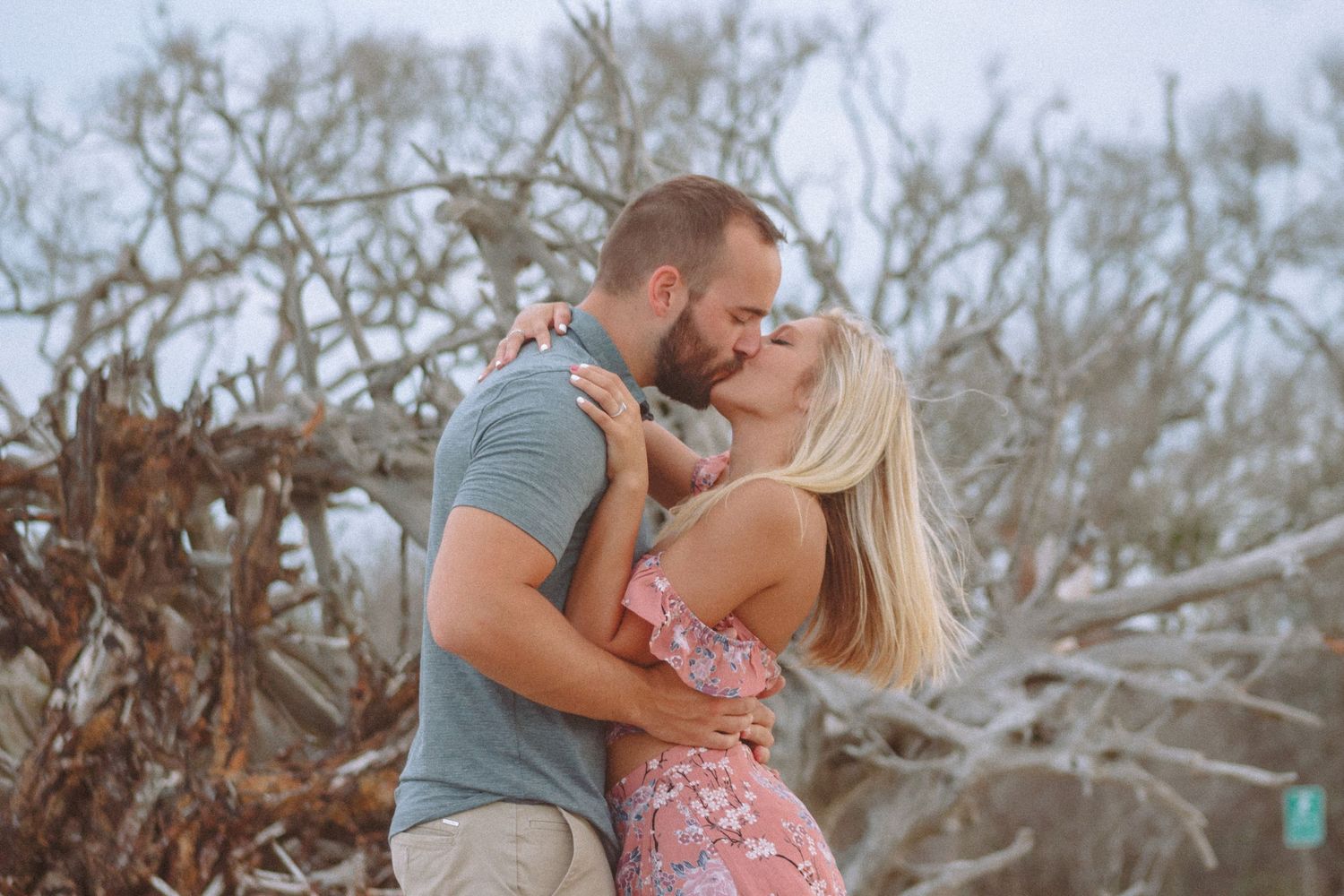 A couple shares a romantic kiss in front of bare twisted branches in an outdoor engagement photo.