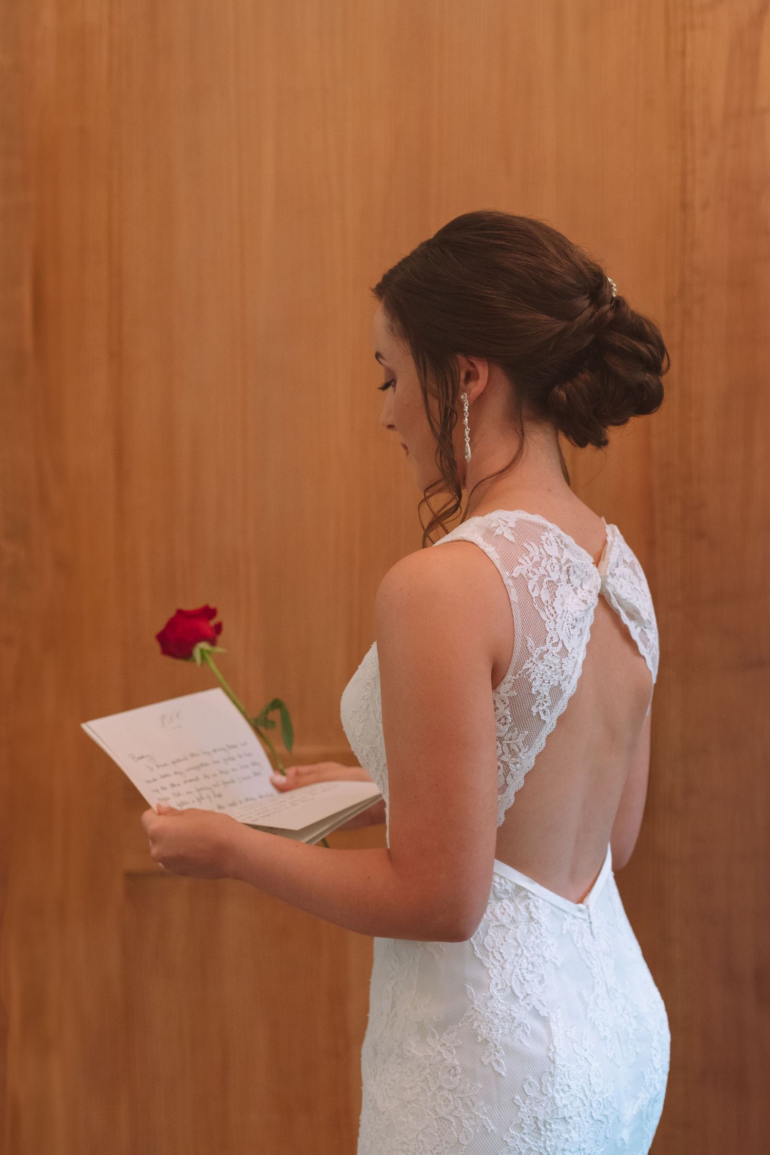 Bride in backless lace wedding dress holds a red rose and reads a letter against a wooden wall.