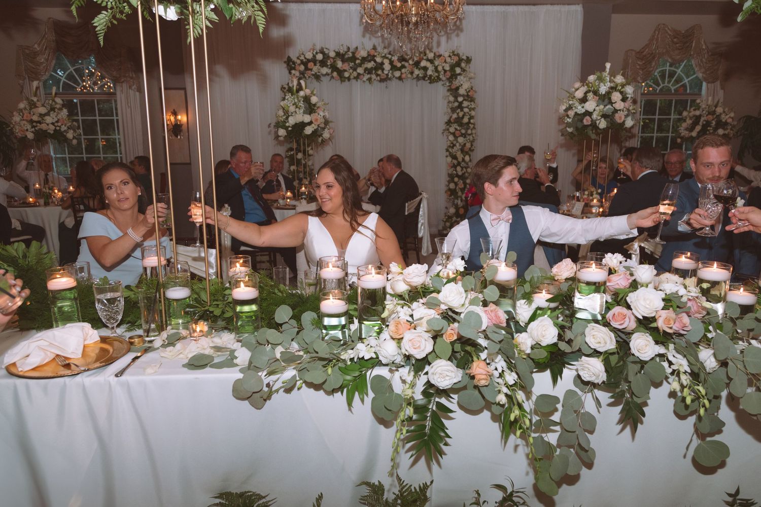 Wedding party raising candles at elegantly decorated head table with floral arrangements.