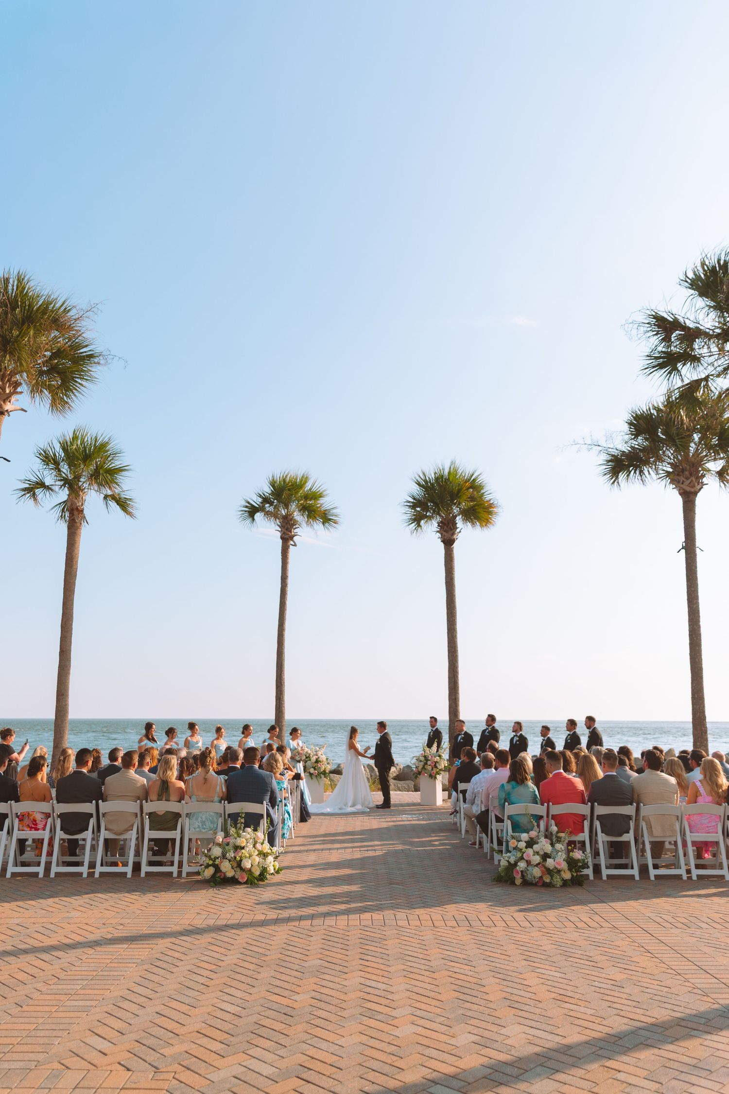 Outdoor beachside wedding ceremony with palm trees and guests seated on white chairs.