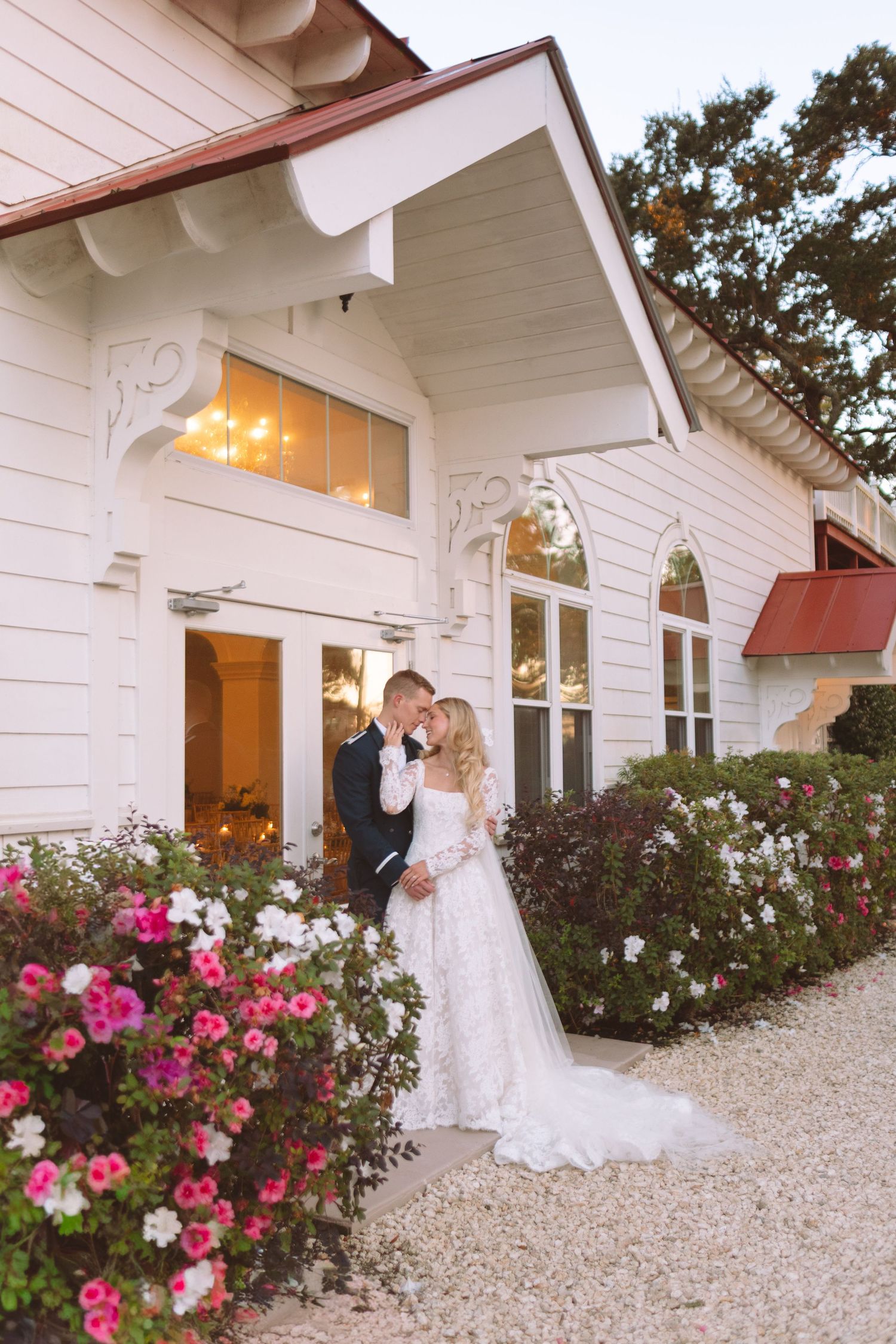 Bride and groom kiss outside a charming white wedding venue surrounded by pink flowers.