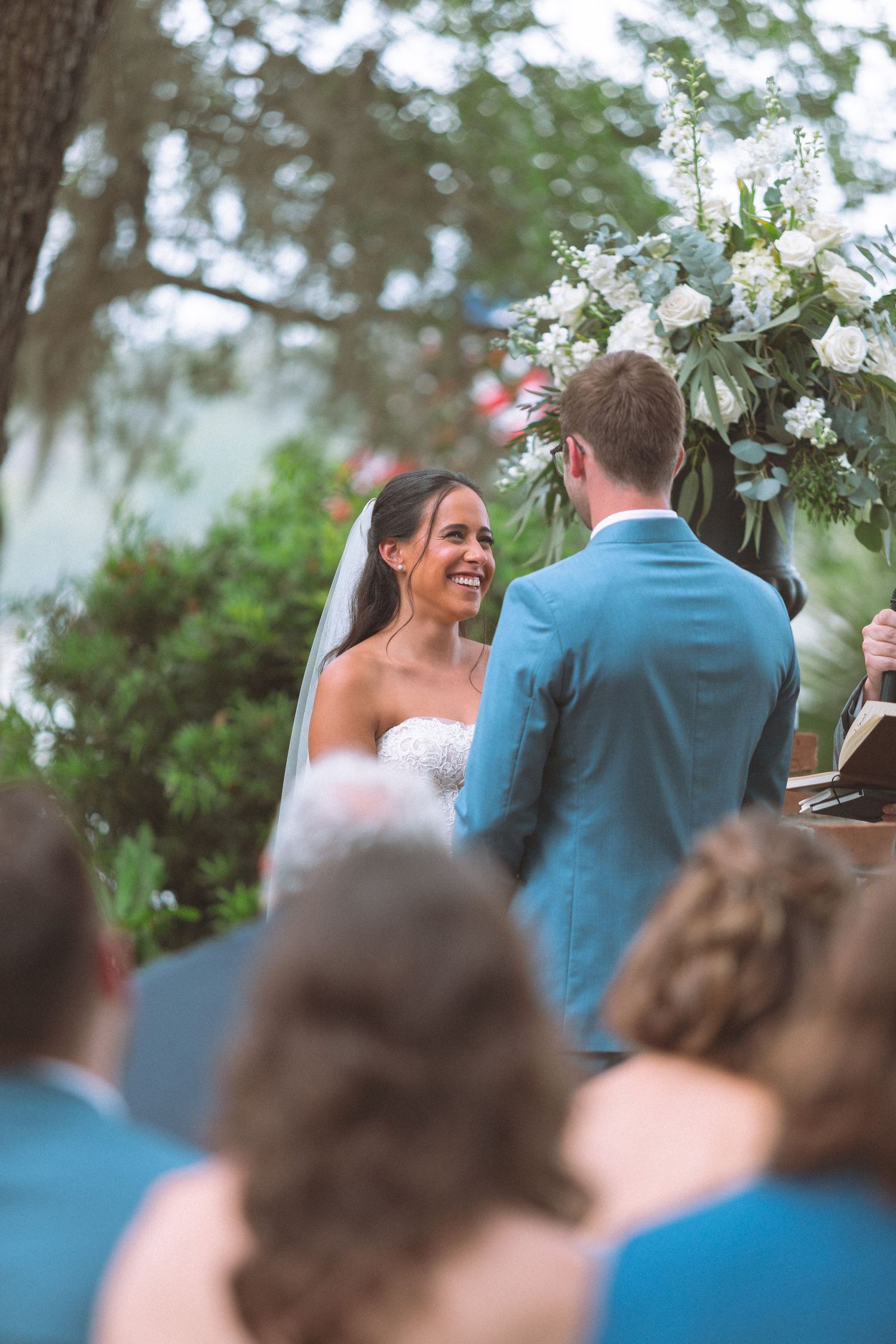Smiling bride in white strapless gown and veil exchanges vows with groom at outdoor wedding ceremony.