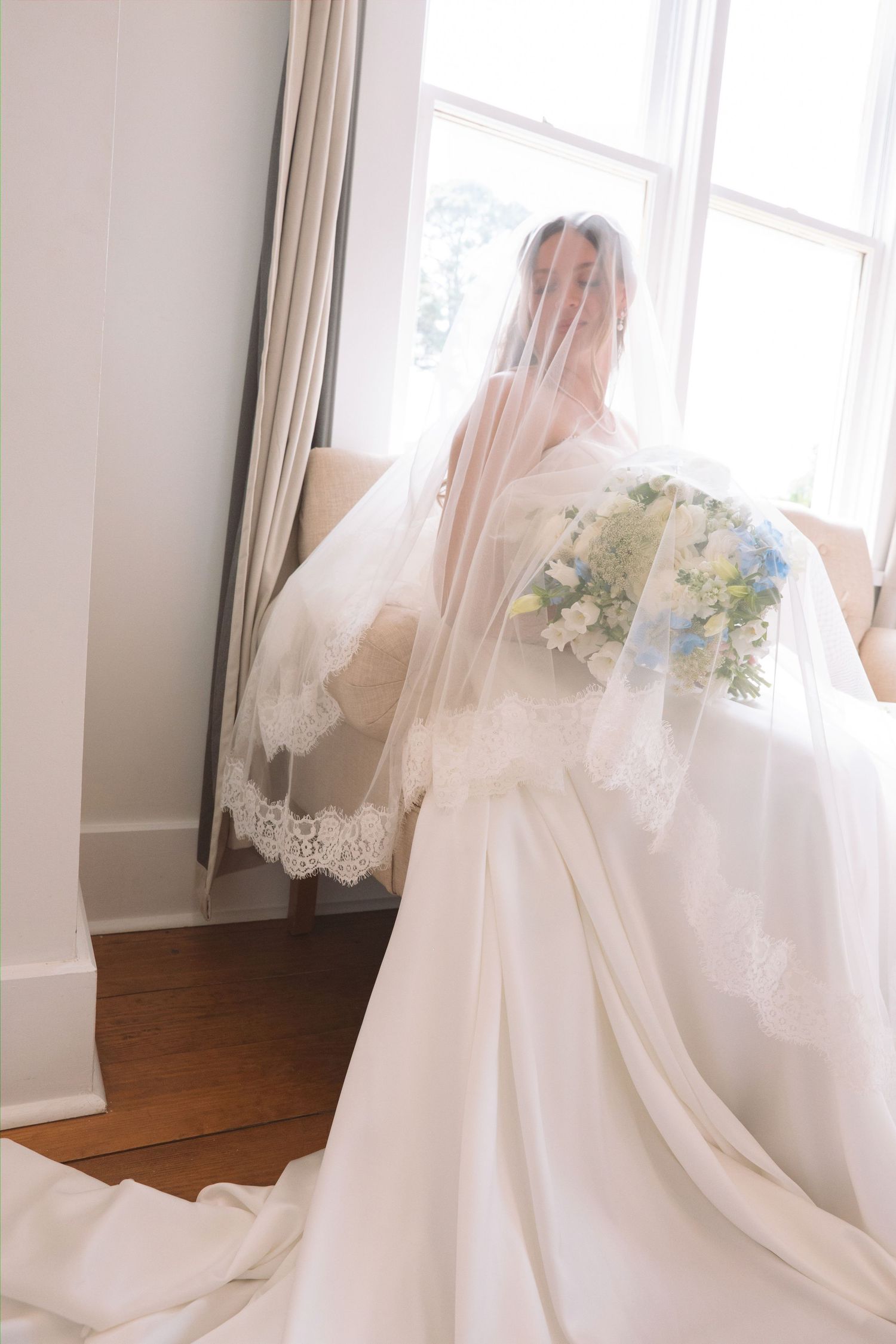 Bride in white ball gown with lace-trimmed cathedral veil holding blue and white bouquet by window.