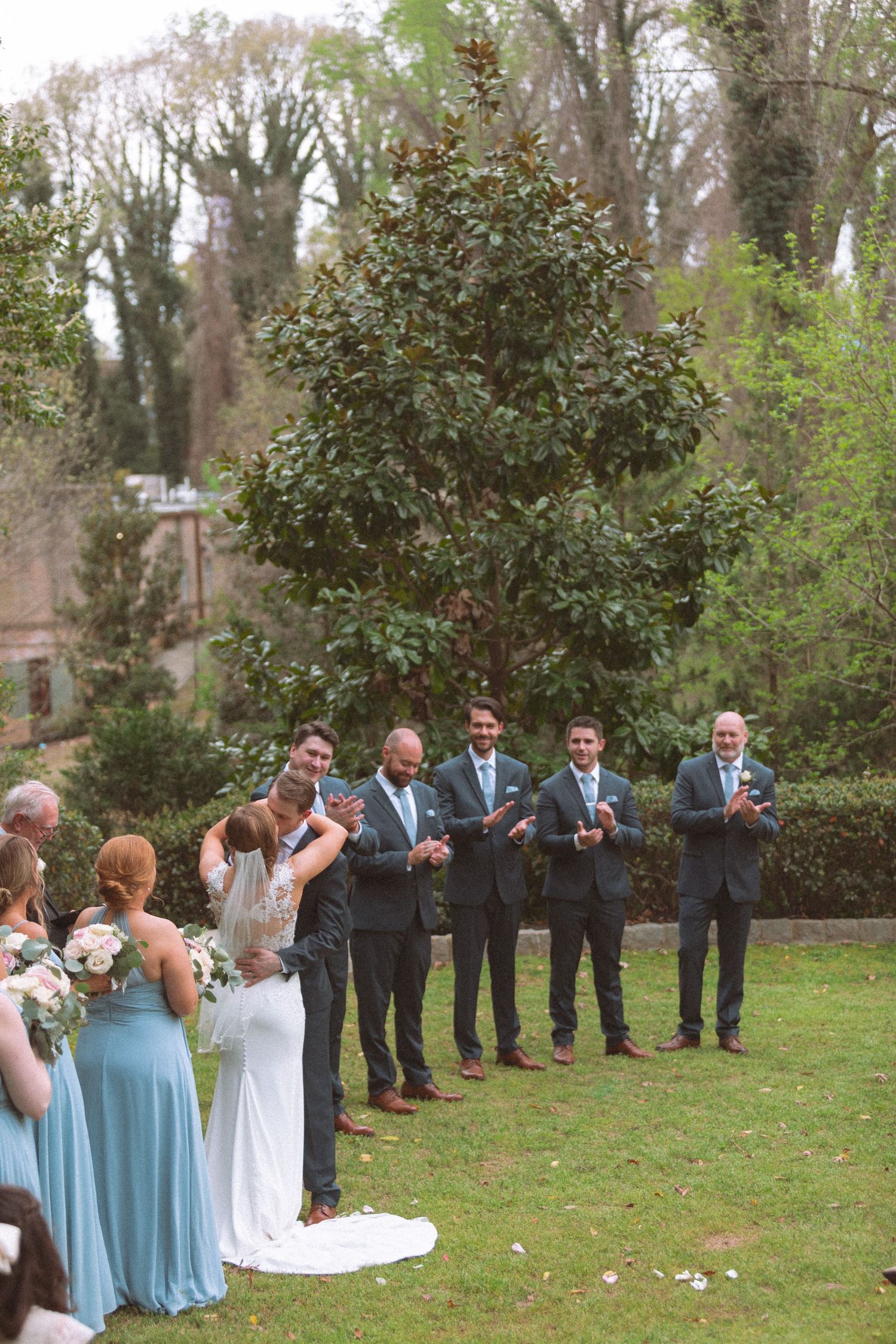 Bride and groom share first kiss at outdoor wedding ceremony while groomsmen in navy suits and bridesmaids in blue applaud.