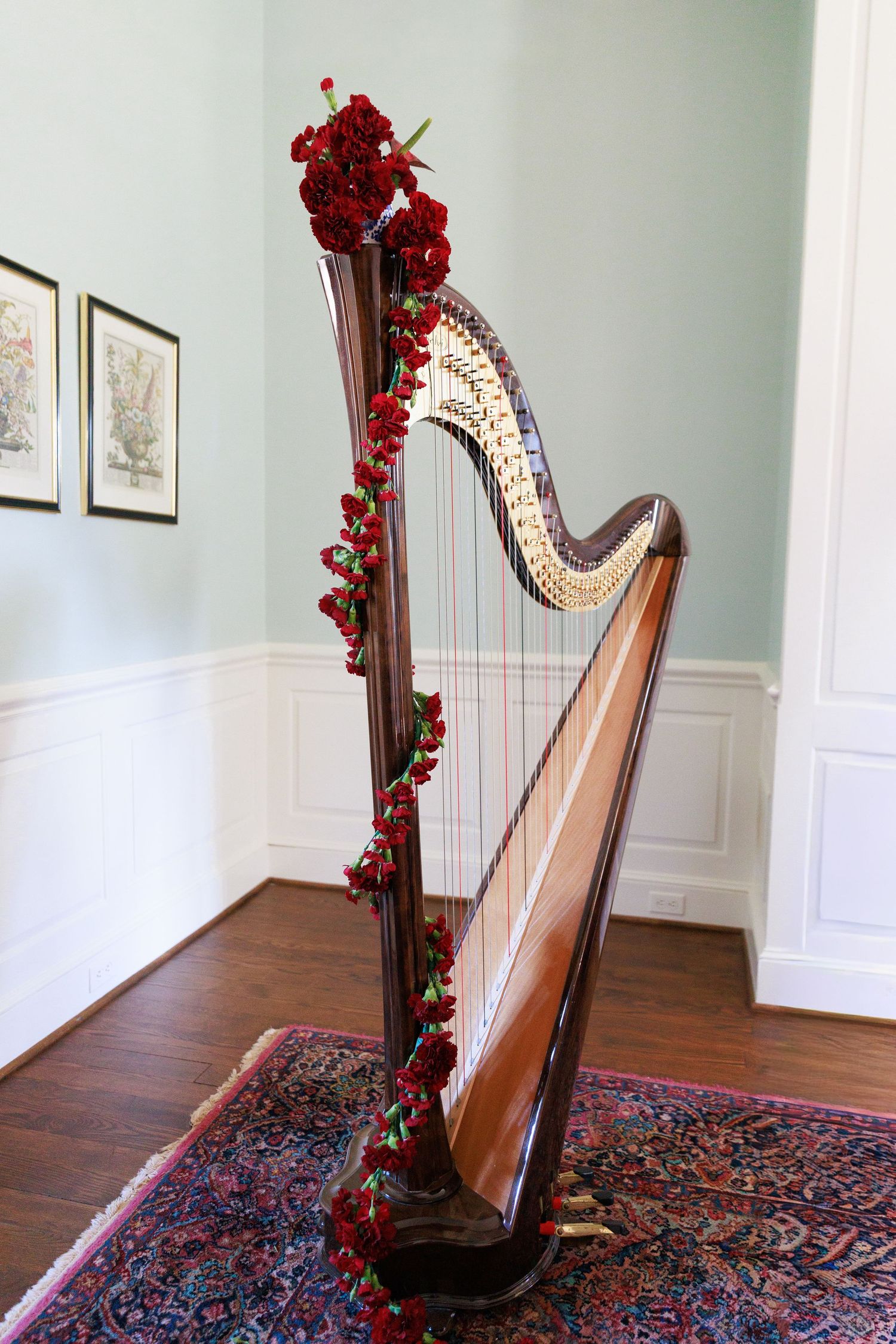 A decorated concert harp adorned with red roses and garland stands on an ornate rug in an elegant room.