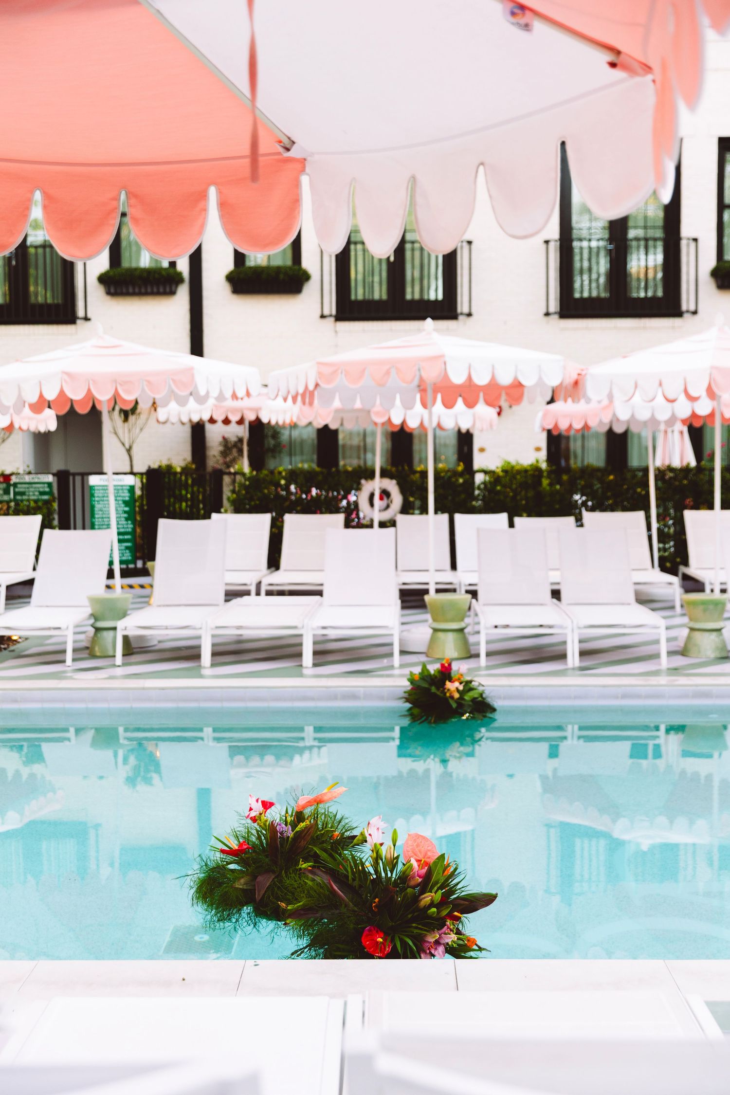 Elegant hotel pool with white lounge chairs and pink fringed umbrellas reflected in turquoise water with tropical floral arrangements.