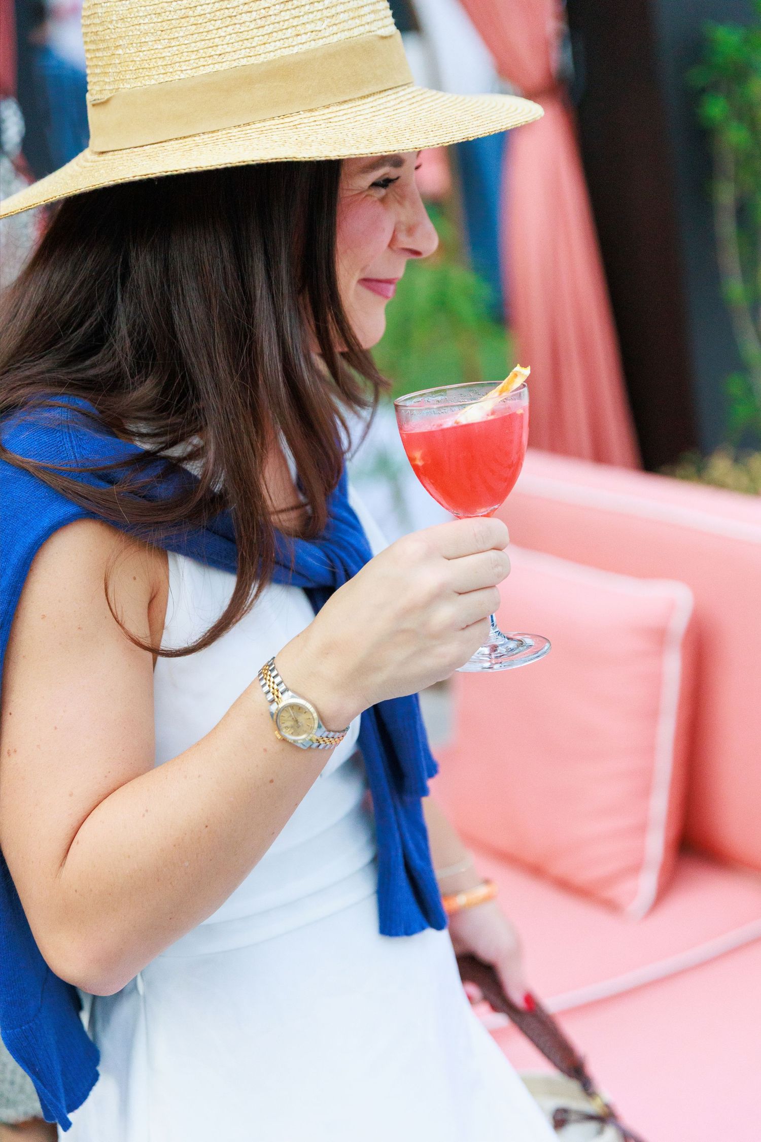 Woman in straw hat and white dress holding a red cocktail in a stemmed glass outdoors near pink seating.