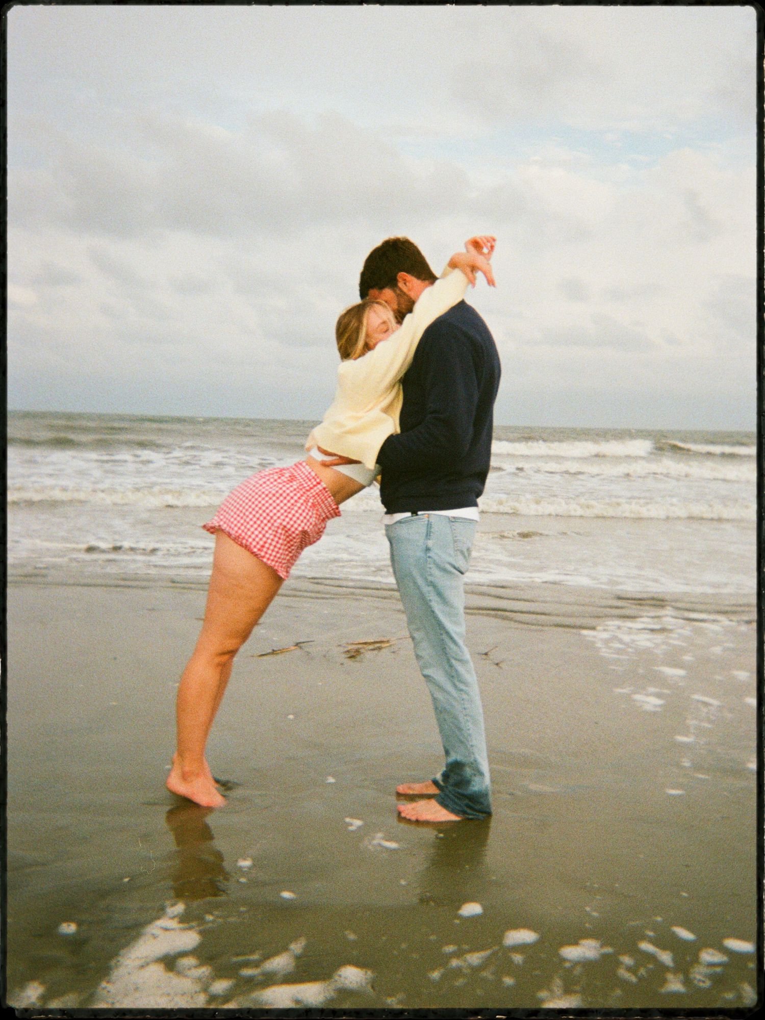 Couple dancing romantically on beach shoreline, woman in red shorts leaning back with waves behind them.