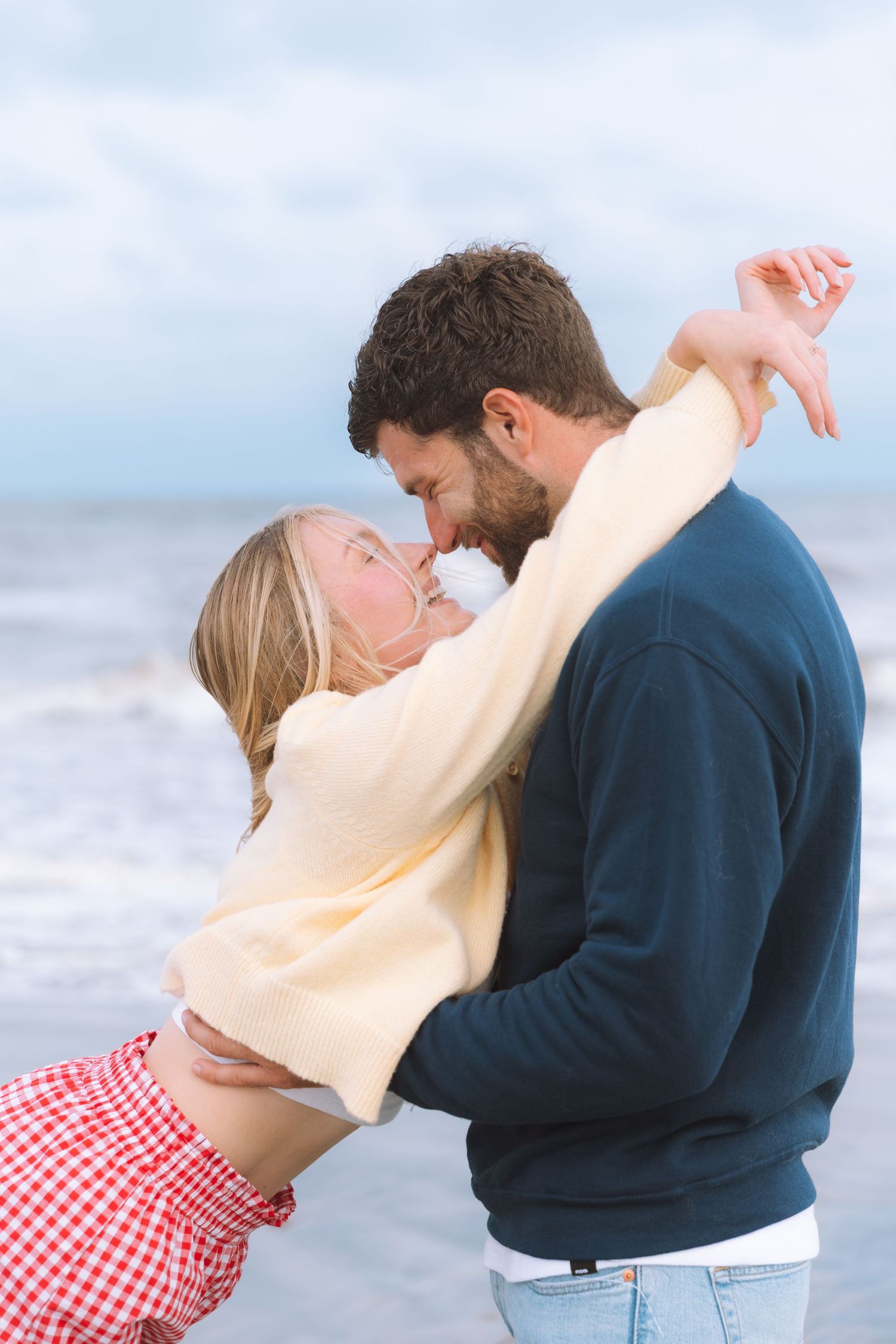 Romantic couple embracing and almost kissing on a cloudy beach, woman in red skirt and cream sweater.