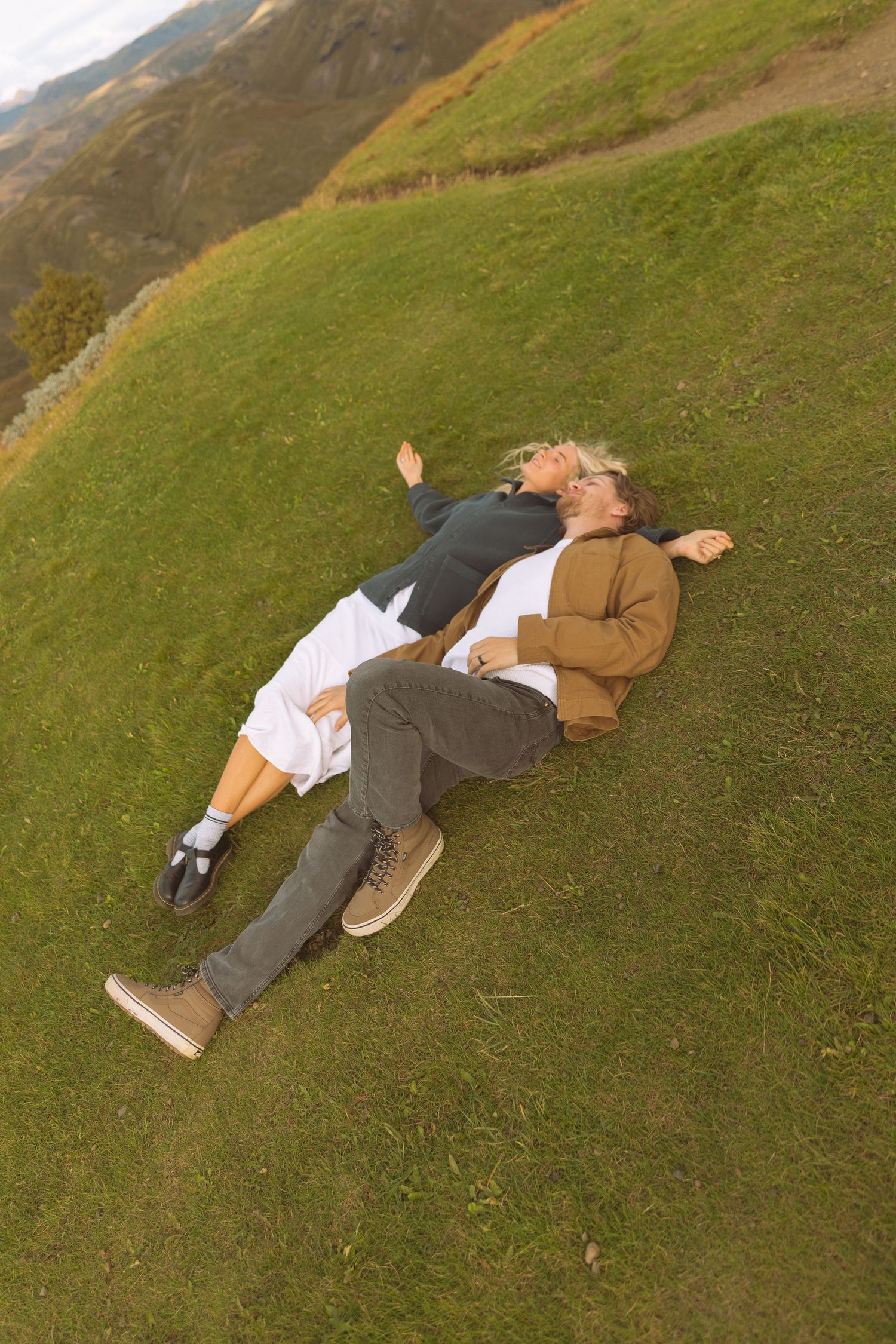 Couple lying together on a lush green hillside with scenic mountain landscape behind them.