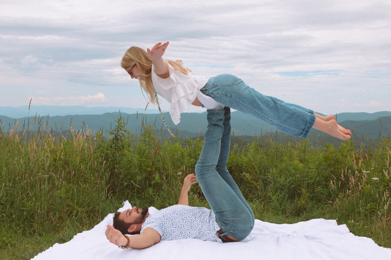 A couple does an acro yoga pose on a white blanket in a green meadow.