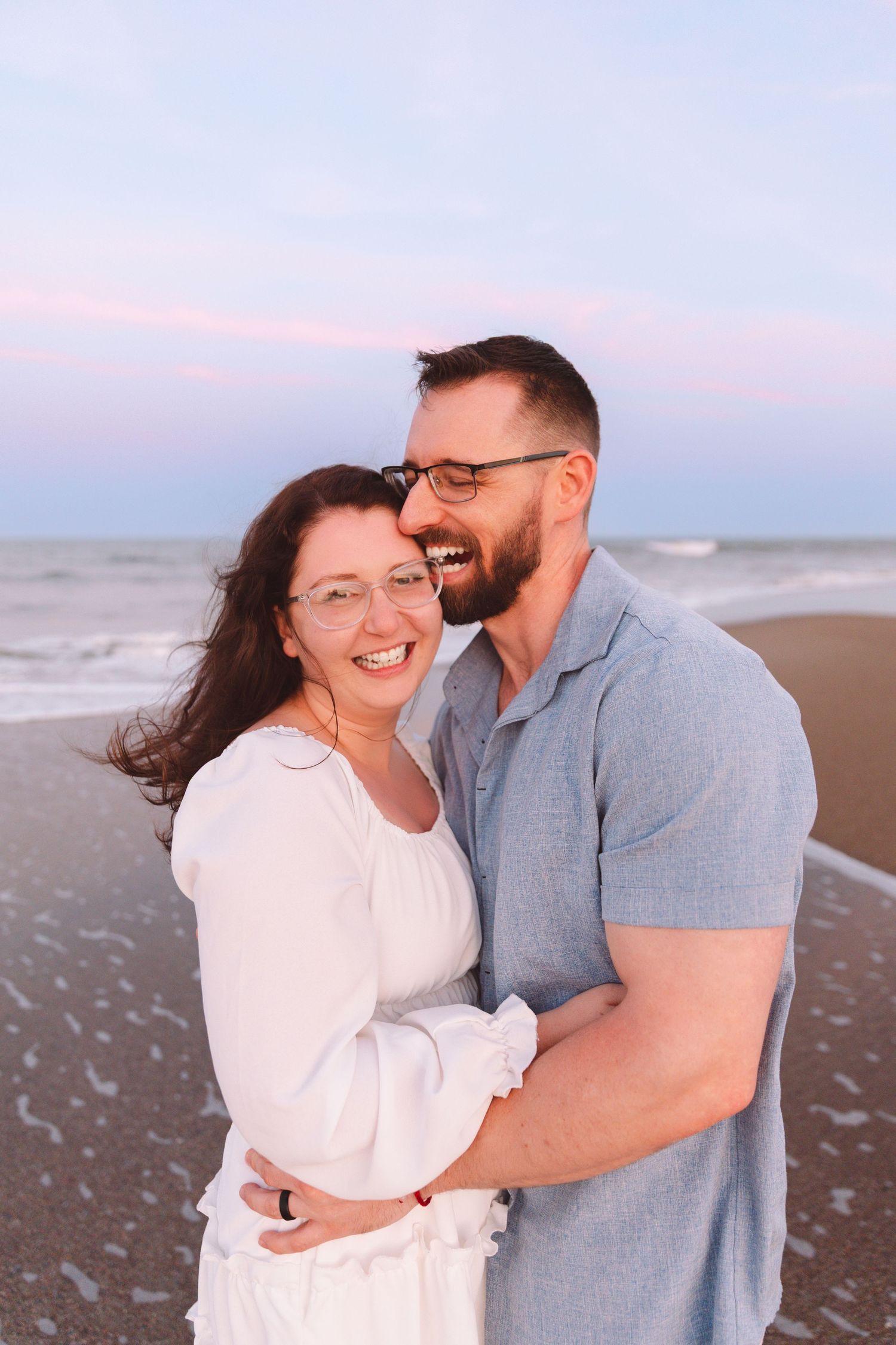 Smiling couple embracing on beach at sunset, woman in white dress and man in blue shirt, both wearing glasses.