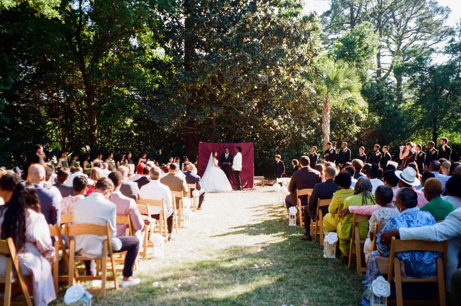Outdoor wedding ceremony with guests seated on wooden chairs facing couple at burgundy floral arch amid trees.