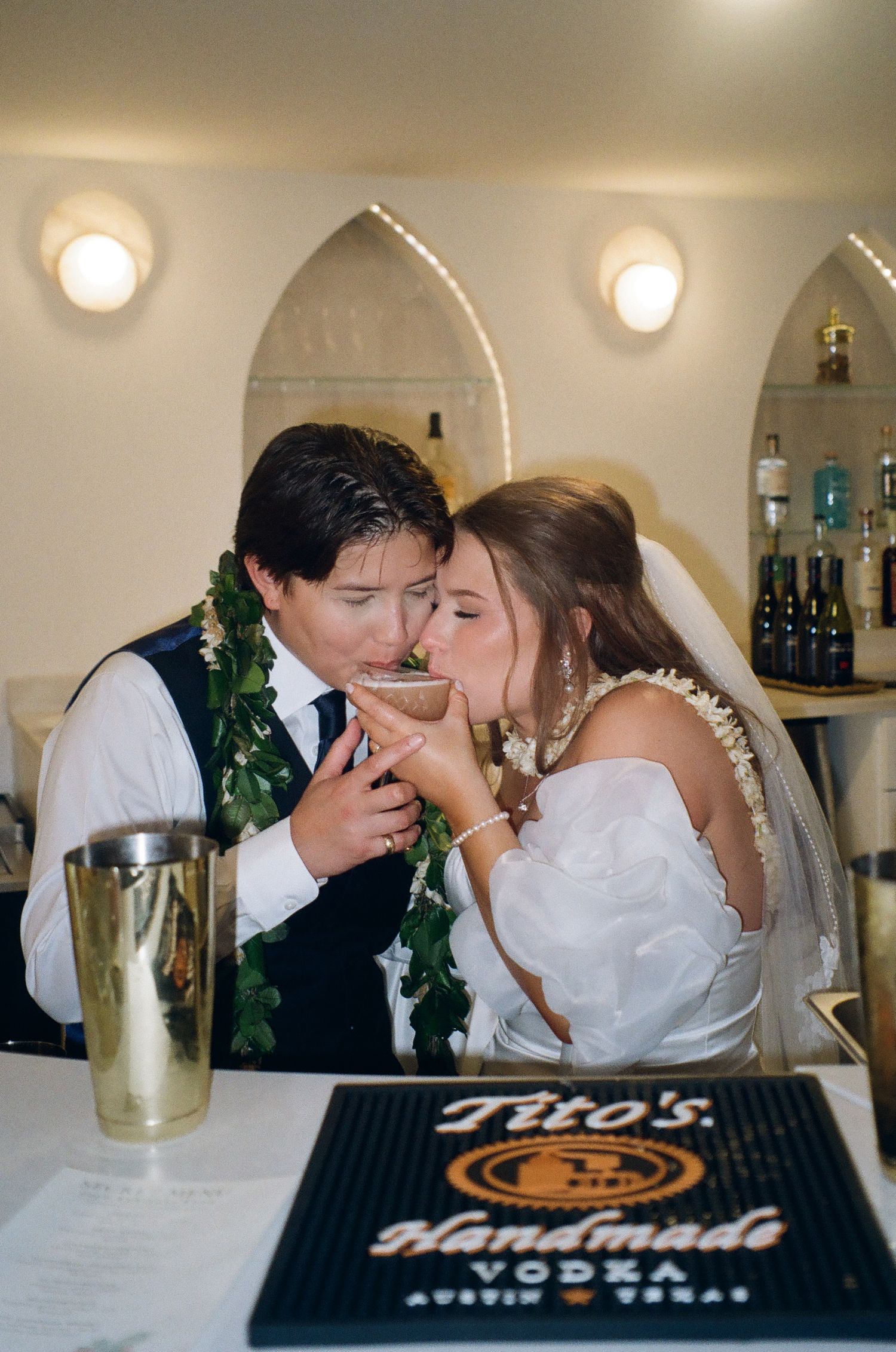 Newlyweds share a drink at a bar with a Tito's Handmade Vodka sign, wearing floral leis at their wedding reception.